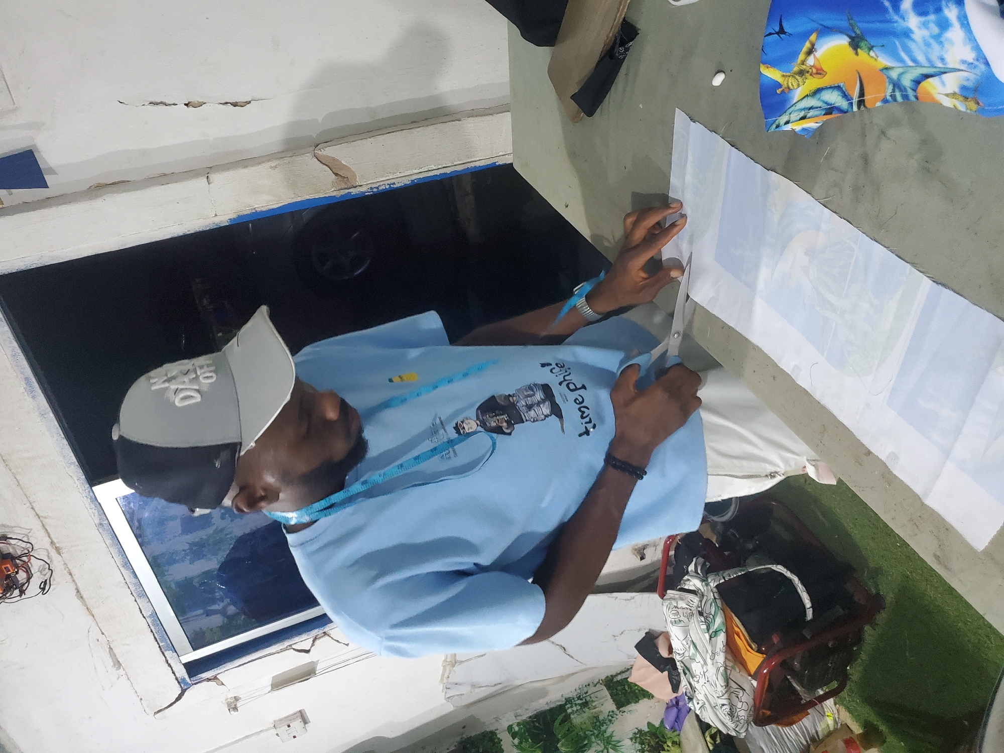 A Black man, wearing a light blue t-shirt, a grey and black baseball cap, and a blue measuring tape around his neck, is shown in an indoor setting. He is in the process of cutting a piece of fabric with a pair of silver scissors. The fabric he is cutting has a colorful pattern featuring dinosaurs. Another piece of fabric, with a similar dinosaur print, is visible in the upper right corner.
The scene appears to be a workspace, possibly a tailoring or sewing area, as indicated by the presence of fabric and sewing tools. The background shows a dark opening, perhaps a window or doorway, with a reflection on its surface. The walls appear to be rough and weathered, with some cracks and peeling paint, suggesting an older building or an unfinished construction.
The man's posture and the way he is holding the scissors and fabric indicate he is focused on his task. The overall impression is one of an individual engaged in a creative or productive activity. The image is shot from a top-down perspective, with the subject oriented vertically within the frame. No specific time of day or weather conditions can be determined from the image.
There is some visible text on the man's t-shirt, though it is partially obscured and difficult to read clearly. It appears to be a graphic design with some lettering.