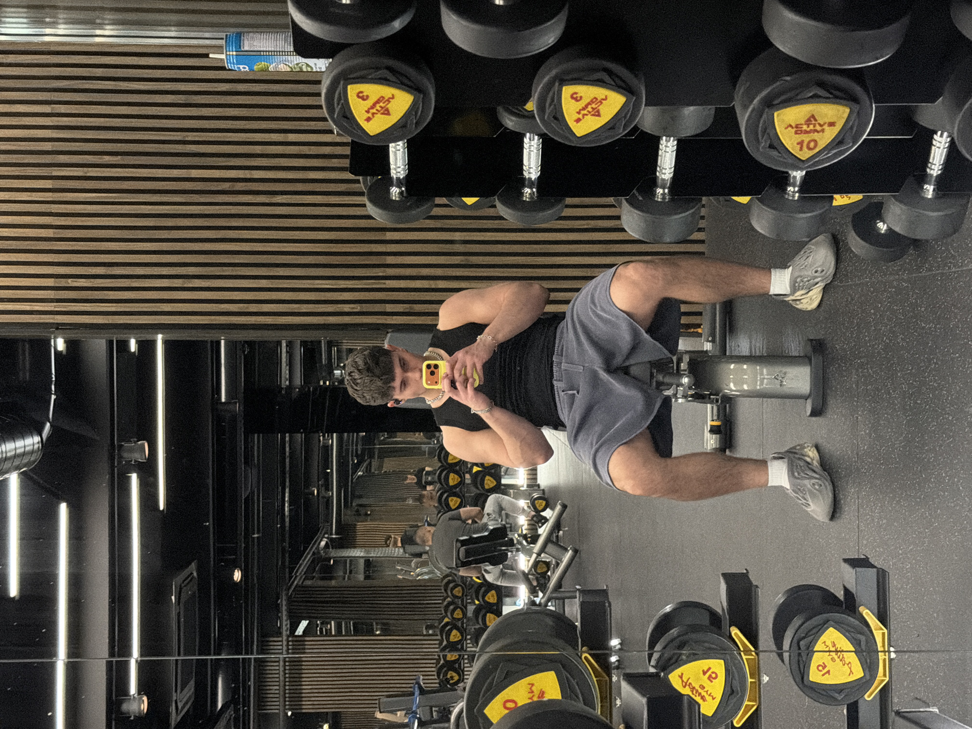 The image shows a man at a gym, presumably in Sofia, Bulgaria. The man, dressed in a black tank top and grey shorts, is sitting on a leg extension machine, looking at his phone. The phone has a yellow case. He is wearing grey sneakers and white socks. He is also wearing a bracelet and a necklace. The gym has black walls with vertical lights, and wooden horizontal slats. A rack of dumbbells, with yellow markings, is visible. Other gym-goers are blurred in the background. A water bottle is placed on a shelf near the ceiling. The scene seems to be reflected in a mirror.