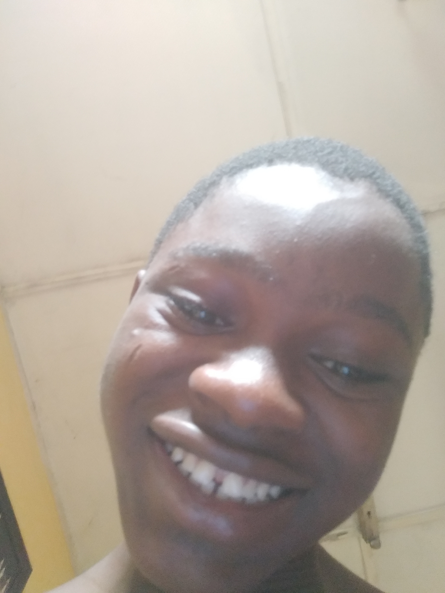 A close-up selfie captures a young person with dark skin and short dark hair, smiling widely at the camera. The individual's face fills most of the frame, conveying a joyful and direct expression. Their broad smile reveals a full set of bright white teeth. Bright specular highlights are visible across the forehead and nose, indicating strong overhead or frontal lighting. Distinct but faint scars or marks are visible on the person's left cheek.
The background is an indoor setting, primarily consisting of a light, off-white colored ceiling or upper wall, with a visible corner line where walls meet. A strip of a lighter yellow or beige wall can be seen on the left side of the frame, and a dark, vertical object is partially visible in the lower-left corner. On the far right, a small, light-colored object, possibly a fixture, is attached to the wall. The image was captured in Shangisha, Nigeria. No visible text is present.