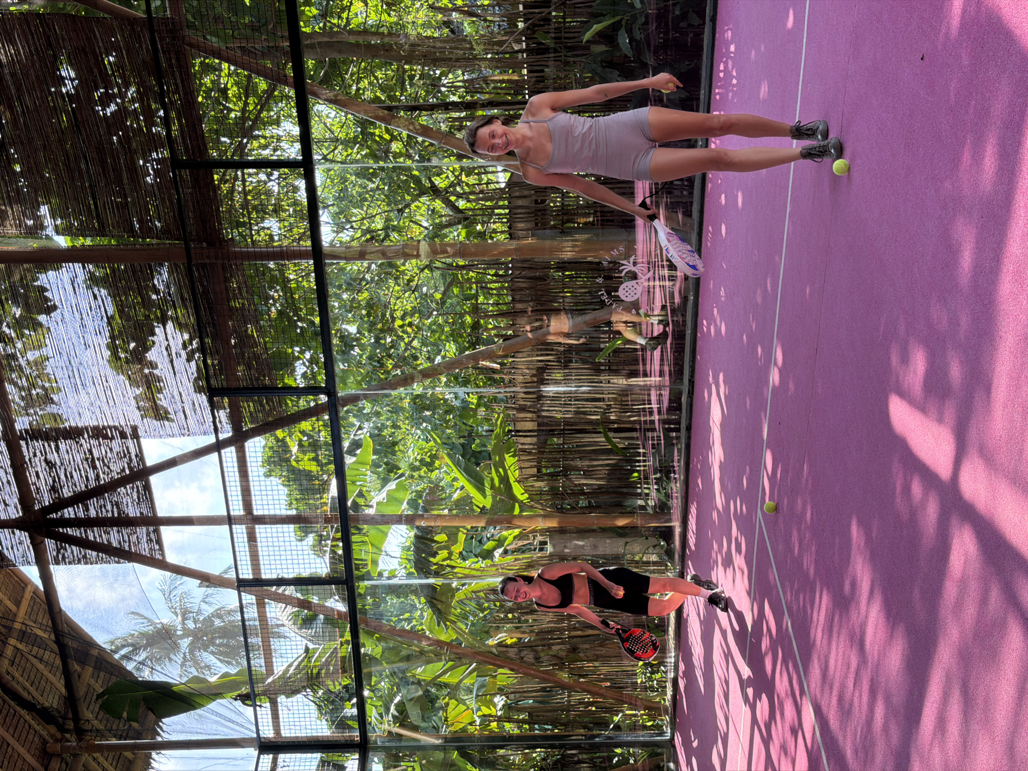 This media file captures two women posing on a vibrant pink padel court, surrounded by lush tropical vegetation in General Luna, Philippines. The image is taken outdoors under bright sunlight, which casts distinct shadows of tree leaves and structures onto the court, indicating midday or early afternoon.
One woman, positioned further back on the court, wears a light grey athletic romper and holds a white padel racket with a yellow ball in her other hand. She is smiling towards the camera. The second woman, closer to the foreground, is dressed in a black sports bra and black athletic shorts, holding a black and red padel racket and a yellow ball. She also smiles at the camera. Both women wear athletic shoes.
The padel court features bright pink surfacing with clear white boundary lines. It is enclosed by transparent walls, part of which includes a metal wire mesh section. Beyond these walls, a dense tropical forest with tall green trees, including what appears to be banana plants and palm trees, forms the backdrop. Above the court, a rustic-looking roof or canopy made of bamboo-like material provides partial shade.
Visible through the transparent court wall, specifically behind the woman in grey, is text that reads "PADEL PALMS" accompanied by a pineapple logo, suggesting the name of the establishment. The overall scene conveys a cheerful and active mood in an idyllic, warm-weather setting.