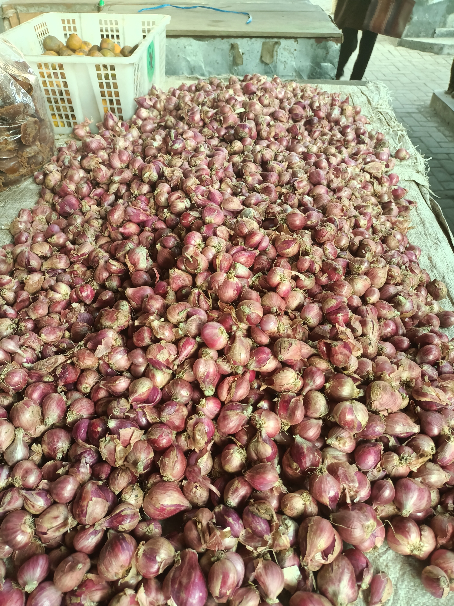 A close-up, high-angle shot shows a large pile of red shallots spread across a light-colored, rough-textured surface, likely a woven mat or tarp. The shallots are mostly a deep pink to reddish-purple color, with some exhibiting lighter, dry papery skins. They are varied in size and shape, with many still showing small dried roots and stems attached.
To the left of the shallots, a clear plastic bag filled with what appears to be palm sugar or a similar dark brown food product is visible. Further to the left and slightly behind the shallots, a white plastic crate contains several rounded, orange-colored fruits, possibly citrus or similar.
In the background, a weathered, light-colored stone wall or structure is partially visible. To the upper right, a person's legs are visible, wearing dark pants or leggings, suggesting they are standing or walking near the scene. The ground in the distance appears to be paved with brick-like stones. The lighting suggests it is daytime, with some shadows cast by the shallots and other objects. The overall scene depicts a market stall or food preparation area, likely in Badas, Indonesia, given the context of red shallots being a common commodity there.