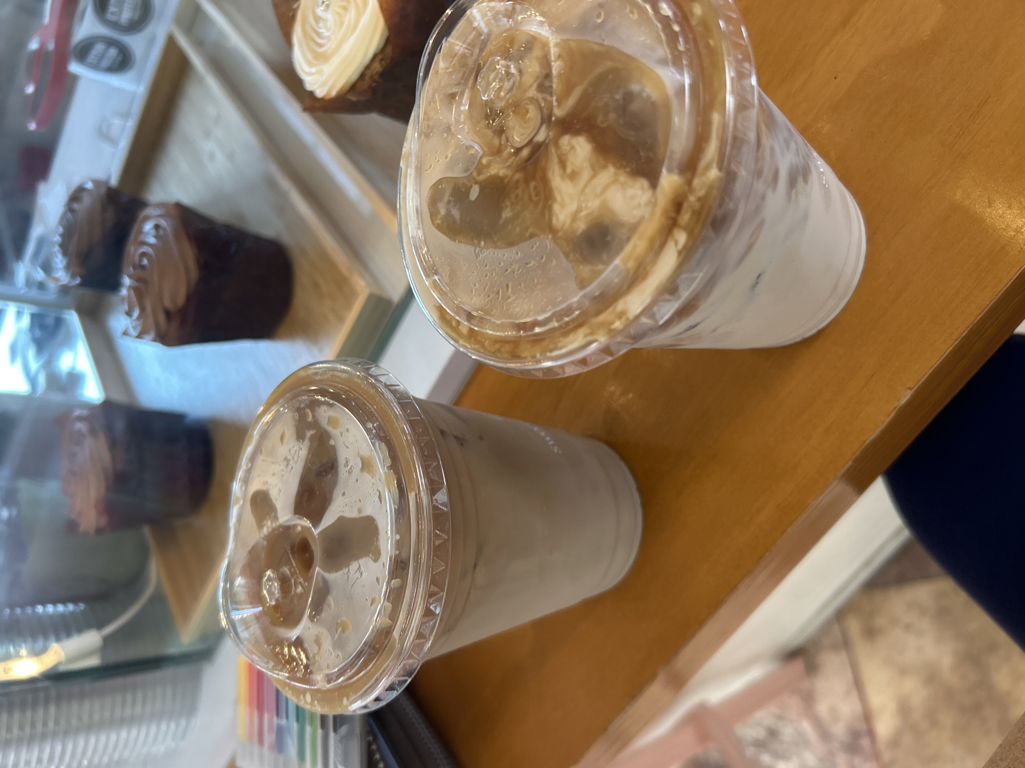 Here is a description of the image:
The photograph shows a cafe or bakery scene. In the foreground, there are two clear plastic cups filled with iced coffee beverages, arranged on a wooden surface, which appears to be a countertop or table. In the background, to the left, there is a display case containing pastries or desserts, specifically what appear to be chocolate pastries. The display case indicates that the setting is a retail establishment, likely a cafe or bakery. The colors in the image are dominated by the browns and tans of the coffee and pastries, and the wood surface. The lighting in the photograph is ambient. A blue article of clothing is slightly visible in the bottom right of the image.