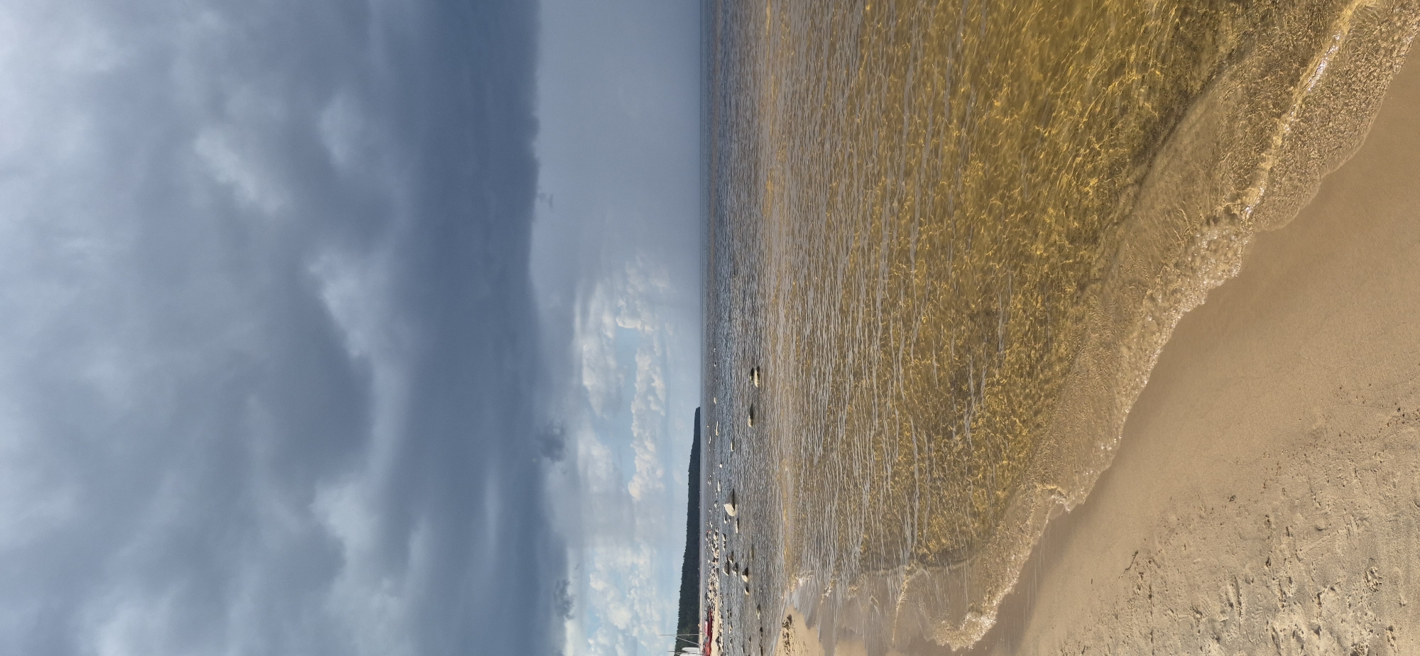 This image depicts a tranquil beach scene in Motornoe, Russia, under a dramatic sky. A sandy shore curves gently into the shallow, amber-colored water. Scattered white stones dot the water's edge and extend further into the sea, some partially submerged. In the distance, a dense line of dark green trees forms the coastline, with a sliver of lighter sky visible above it. The sky itself is a focal point, dominated by heavy, dark grey clouds that create a sense of impending weather. A faint, lighter patch in the sky suggests a break in the clouds or the distant onset of rain. No people are visible in the image.