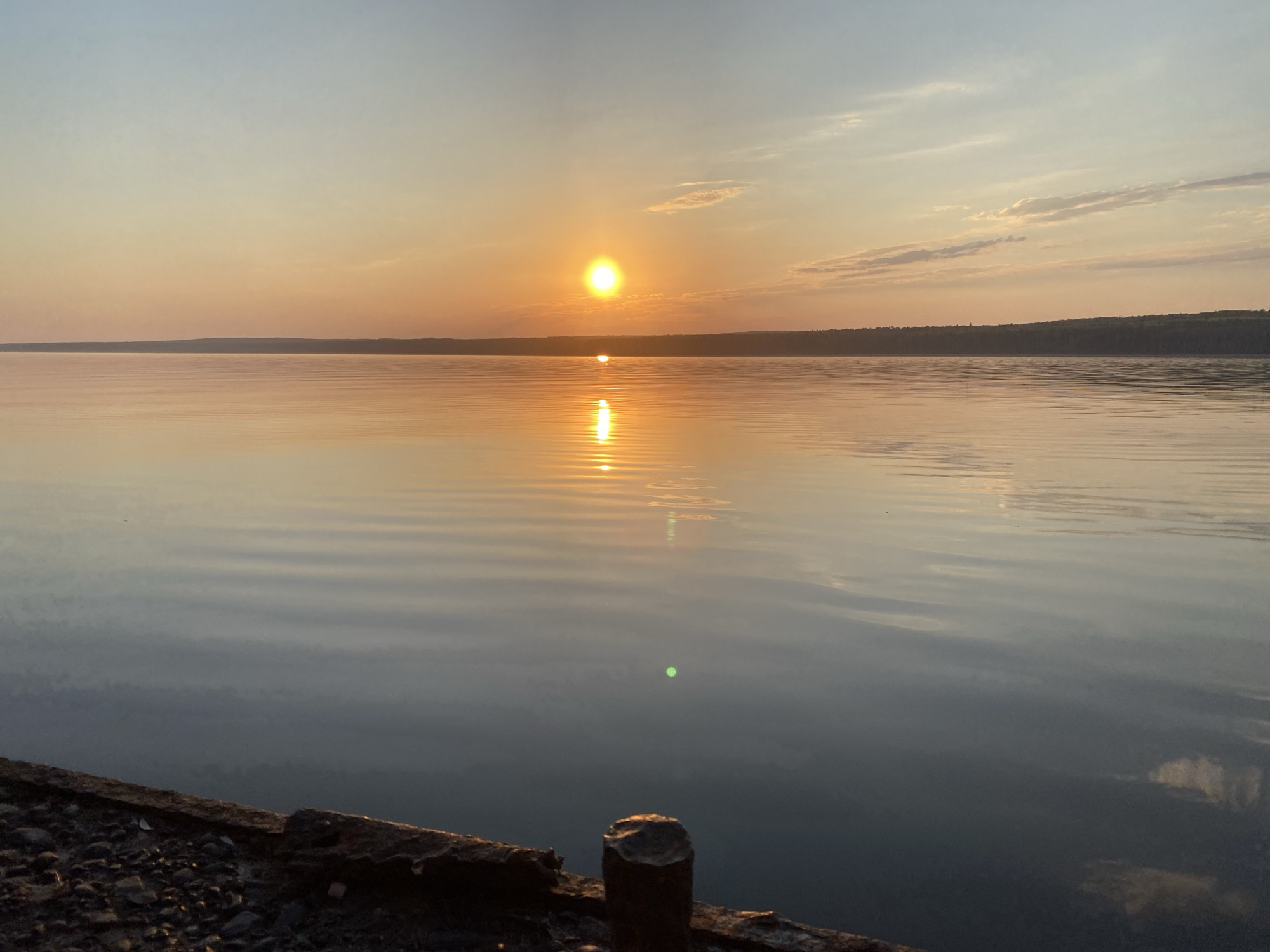 The image captures a sunset over a body of water, possibly a reservoir or a wide river, in what appears to be the city of Chaikovsky, Russia. The sun is positioned low on the horizon, casting a bright reflection on the water's surface. The sky transitions from a lighter hue near the top to a warm, orange color closer to the horizon. In the distance, a dark, forested landmass is visible. The foreground shows a concrete structure with what appears to be a piling extending from the rocks, possibly a dock or part of an embankment. The water has gentle ripples, suggesting calm conditions. There are no people visible in the scene.