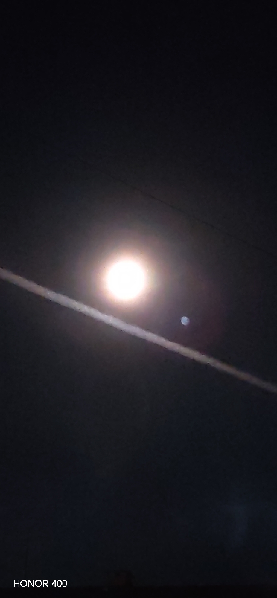 A photograph taken with an Honor 400 mobile device shows the moon in the night sky. The moon is a bright, circular light source in the center of the image, with a thin, horizontal contrail running across the moon. A smaller light appears to be captured near the right side of the moon. The sky is otherwise dark and clear. There is an indistinguishable dark mass at the bottom of the image. The location is unknown but speculated to be in Noviy, Russia. The time of capture is not provided. The image contains no people or interactions.