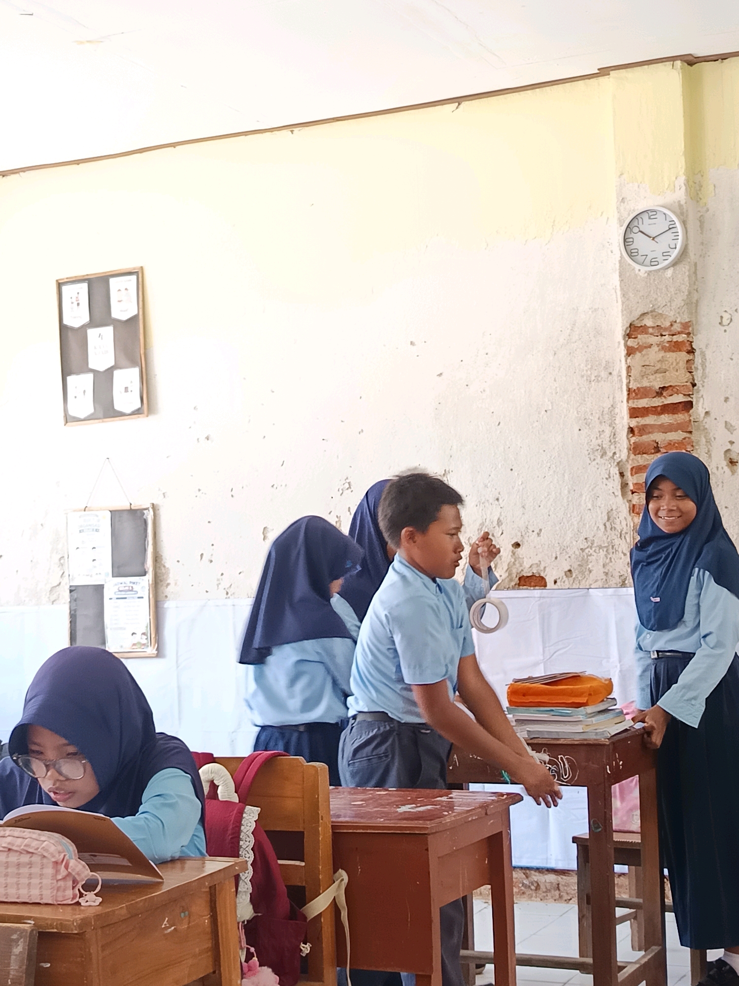 The image depicts a classroom scene in Labuhan, Indonesia, focusing on several students. In the foreground, a young girl wearing a hijab and glasses is seated at a wooden desk, engrossed in reading a book. A backpack hangs on the back of her chair. To the right, two students are interacting near a table laden with books. A boy in a light blue shirt and dark pants is holding a roll of tape, appearing to be involved in an activity. Beside him, a girl in a dark blue hijab and light blue uniform smiles as she observes. In the background, a clock is visible on the wall, and two framed displays hang on the wall, one with multiple smaller pictures and another with text. The room appears to be a classroom with aged walls and furniture.
