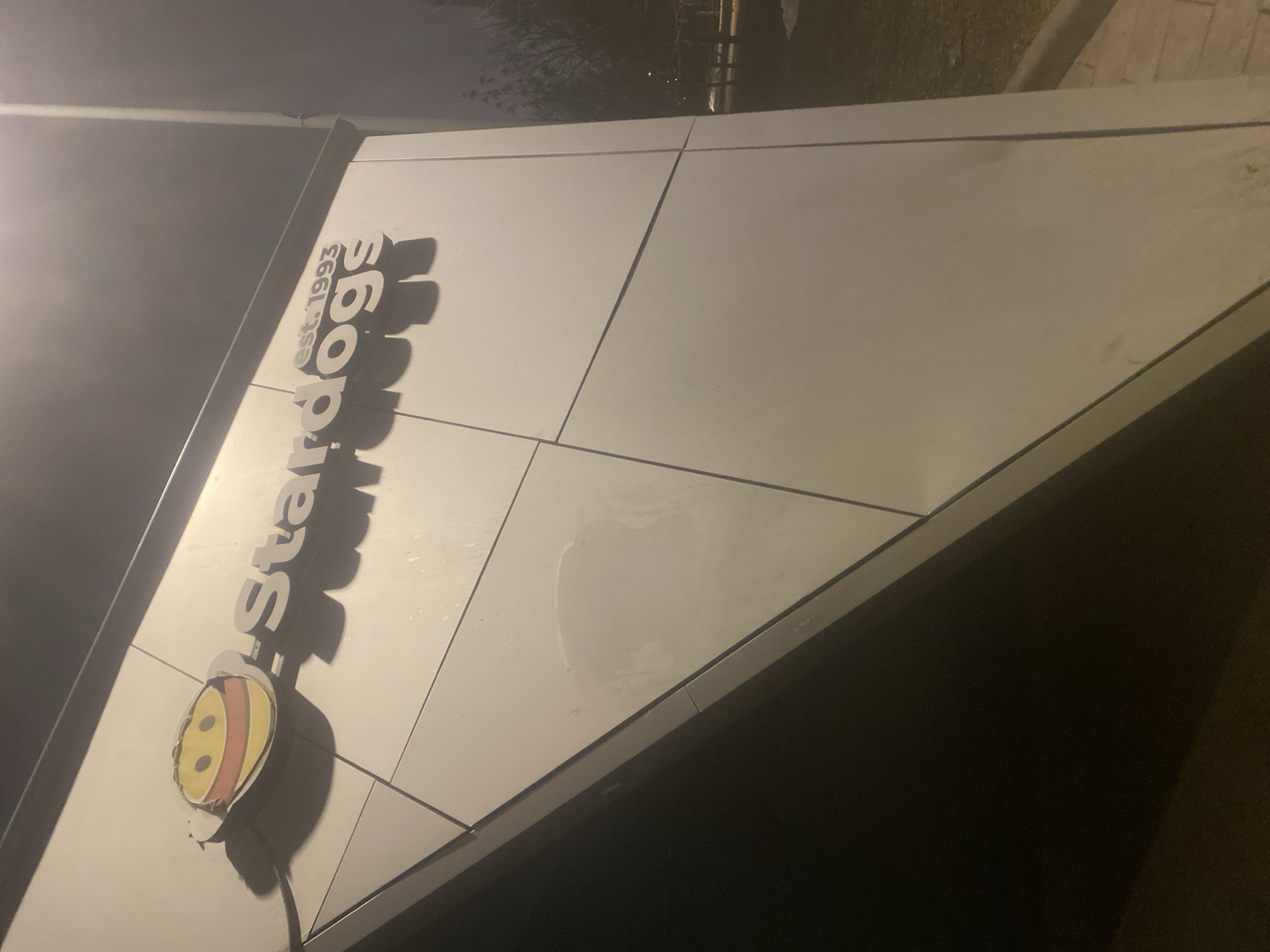The image shows a building sign for "Star-doggs," established in 1993, with a cartoon dog logo. The location is in an unknown city. The sign is made of light-colored panels with dark-colored trim. There is some streaking on the panels. The photo was taken at night, with some artificial light illuminating the sign. The immediate surroundings are out of focus, but appear to be adjacent to a paved area.
