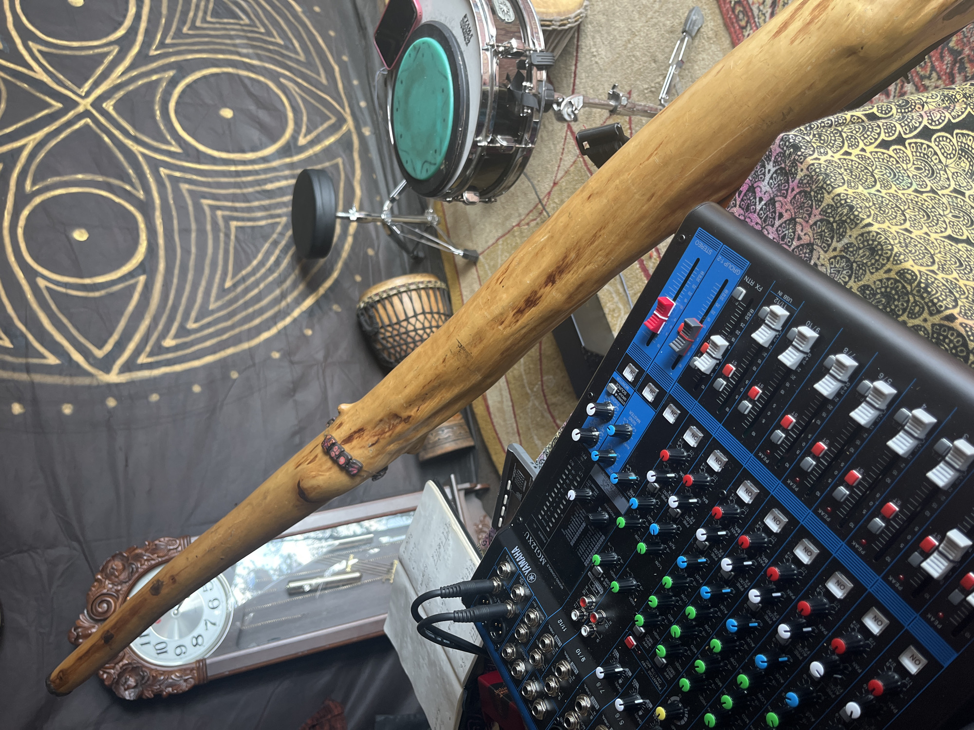 The image depicts a scene with various musical instruments and audio equipment, possibly set up in a room or studio. A prominent, long wooden didgeridoo is positioned diagonally across the frame, leaning against a black fabric backdrop decorated with a gold mandala-like pattern. In the foreground, a black Yamaha MG12XU mixing console is laid out, with numerous knobs, sliders, and buttons visible. Two black audio cables are plugged into the console.
Behind the mixing console and to the left, a grandfather clock with a white face and Roman numerals is visible. Next to the clock, an open notebook with handwritten text rests on a surface.
Further back and to the right, a snare drum with a green practice pad on top is set up on a stand. Adjacent to the snare drum, a small hand drum, possibly a djembe, is also present. A black stool is positioned behind the snare drum. A pink smartphone is resting on the snare drum.
The overall environment appears to be an indoor space, with a patterned rug partially visible on the floor. The lighting is somewhat dim, suggesting an indoor setting or a deliberate atmospheric choice for the setup. There are no people visible in the image.