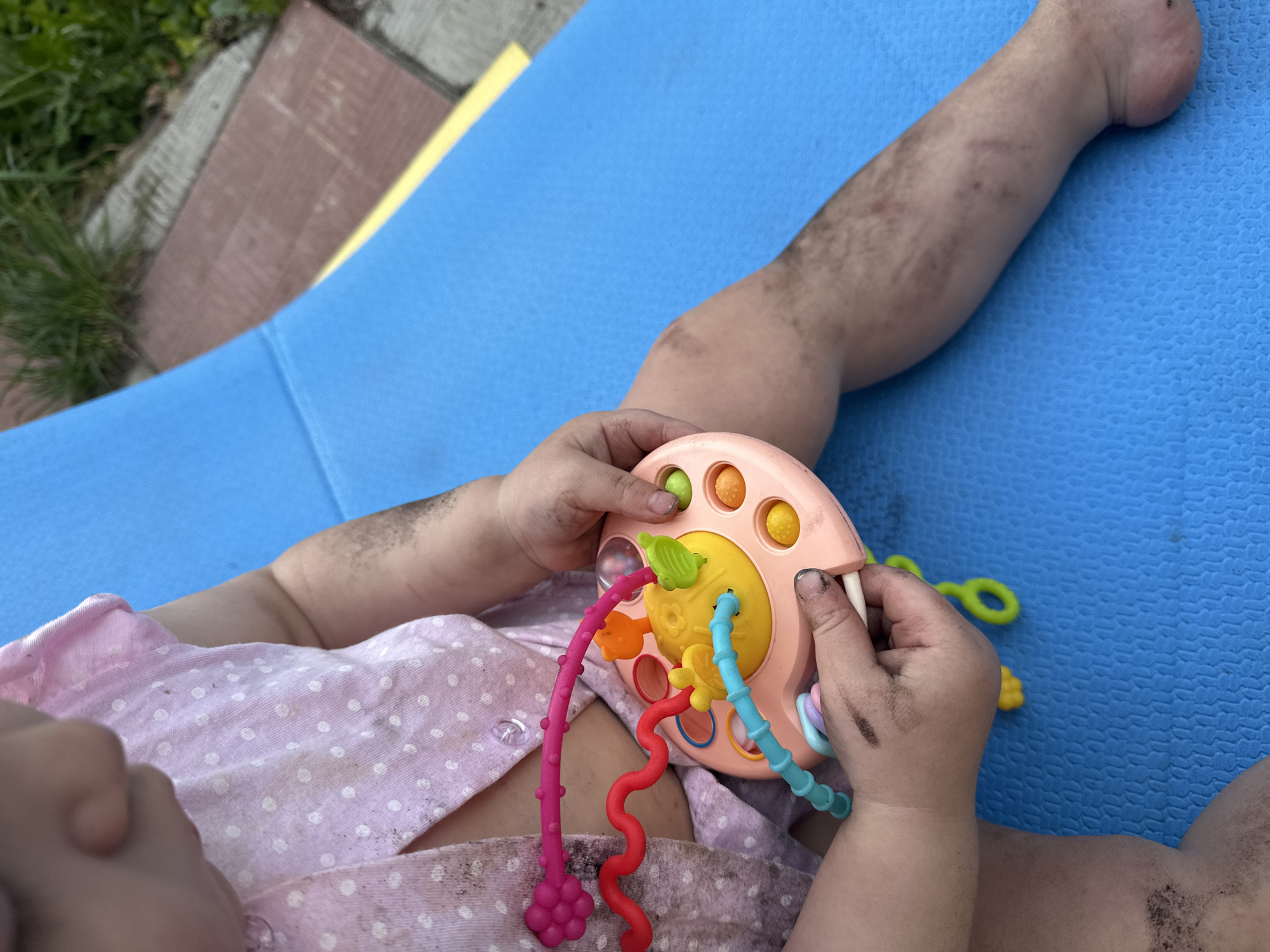 A child is lying on a blue and yellow surface, possibly an outdoor mat or lounger. The child is wearing a pink and white polka-dot shirt and appears to be holding a colorful toy with various shapes and textured strands. The child's hands, arms, and legs are visibly covered in what appears to be dirt or dark residue. The scene is outdoors, with grass and paving visible in the background. There is no indication of the location being Nizhny Novgorod, Russia.