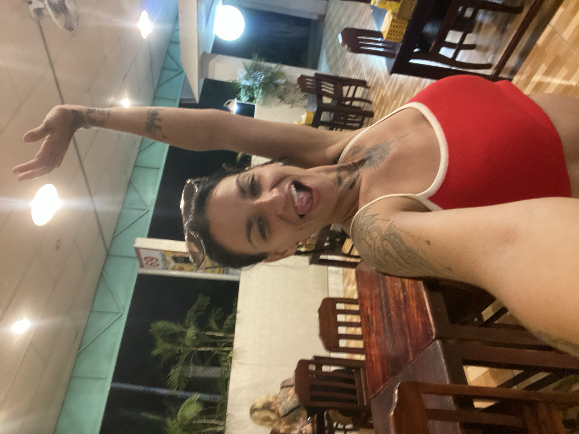 This is a selfie taken by a woman with dark hair, wearing a red and white tank top. She is in an indoor restaurant or cafe, with wooden tables and chairs visible. The scene appears to be set at night, indicated by the artificial lighting and the darkness outside, which is partially visible through an open area with palm trees. The woman is smiling and sticking her tongue out, conveying a playful and energetic mood. She has tattoos on her arms and shoulder. A sign with Vietnamese text and the number "22" is visible in the background, offering a potential clue to the establishment's identity or a location marker. The flooring is light-colored wood. The overall atmosphere is casual and lively.