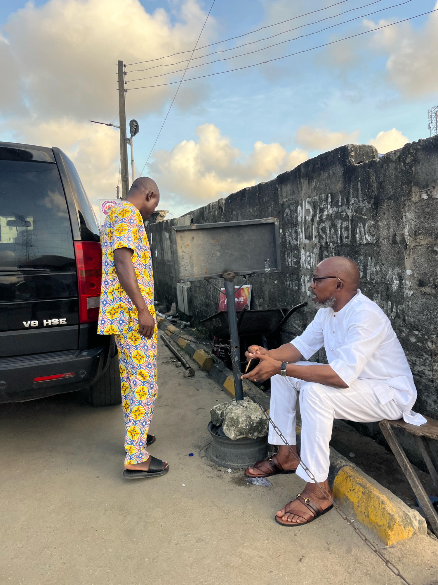 In Ado, Nigeria, on a street with a textured concrete ground, two men are engaged in conversation. The man on the left, dressed in a vibrant yellow and blue patterned traditional outfit, stands facing the man on the right. He wears black sandals. The man on the right, seated on a wooden bench next to a rough, grey concrete wall, is clad in a white traditional outfit and brown sandals. A metal chain links his ankle to a piece of metal on the ground. Behind him, a wheelbarrow is visible. To the left of the men, a black SUV with "V8 HSE" emblazoned on its rear is parked. The background features a cloudy sky with indications of power lines and a utility pole.