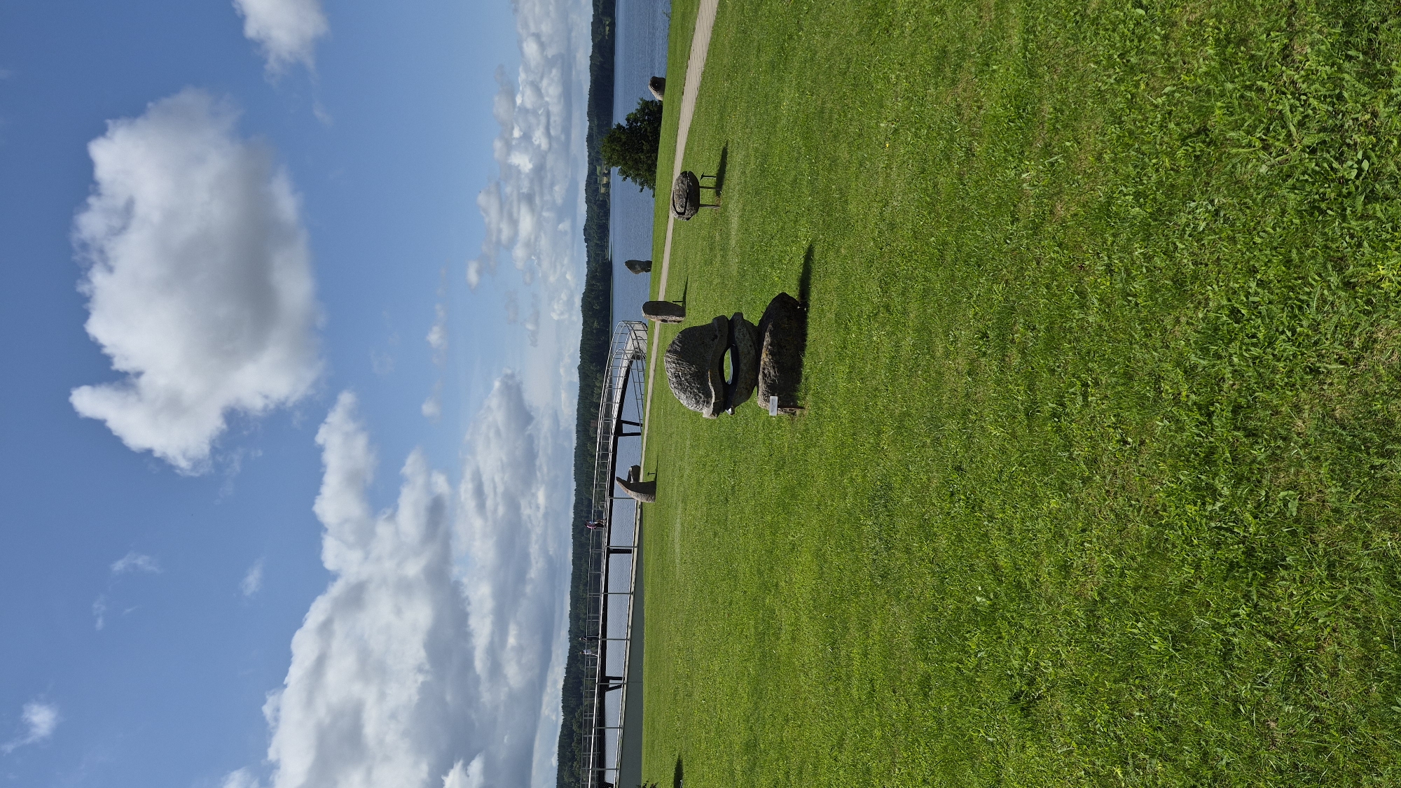 The image captures an outdoor scene in Zarasai, Lithuania, likely at the Zarasai Observation Deck, a popular tourist attraction situated on the shores of Lake Zarasai. The foreground is dominated by a vibrant green, well-maintained lawn, speckled with various rock sculptures of abstract and organic forms. One prominent sculpture in the mid-ground features two round, carved stones stacked upon each other, resembling an open-mouthed creature.
In the background, a wooden pedestrian bridge with a curved design extends over the placid, blue waters of Lake Zarasai. Several indistinct figures, appearing to be people, are visible walking on the bridge, suggesting moderate visitor activity. The lake is fringed by a lush, green treeline, indicative of the natural landscape surrounding the town. The sky above is a clear blue, punctuated by fluffy white clouds, casting soft shadows on the scene. The overall atmosphere is serene and picturesque, characteristic of a summer day in a lakeside park.