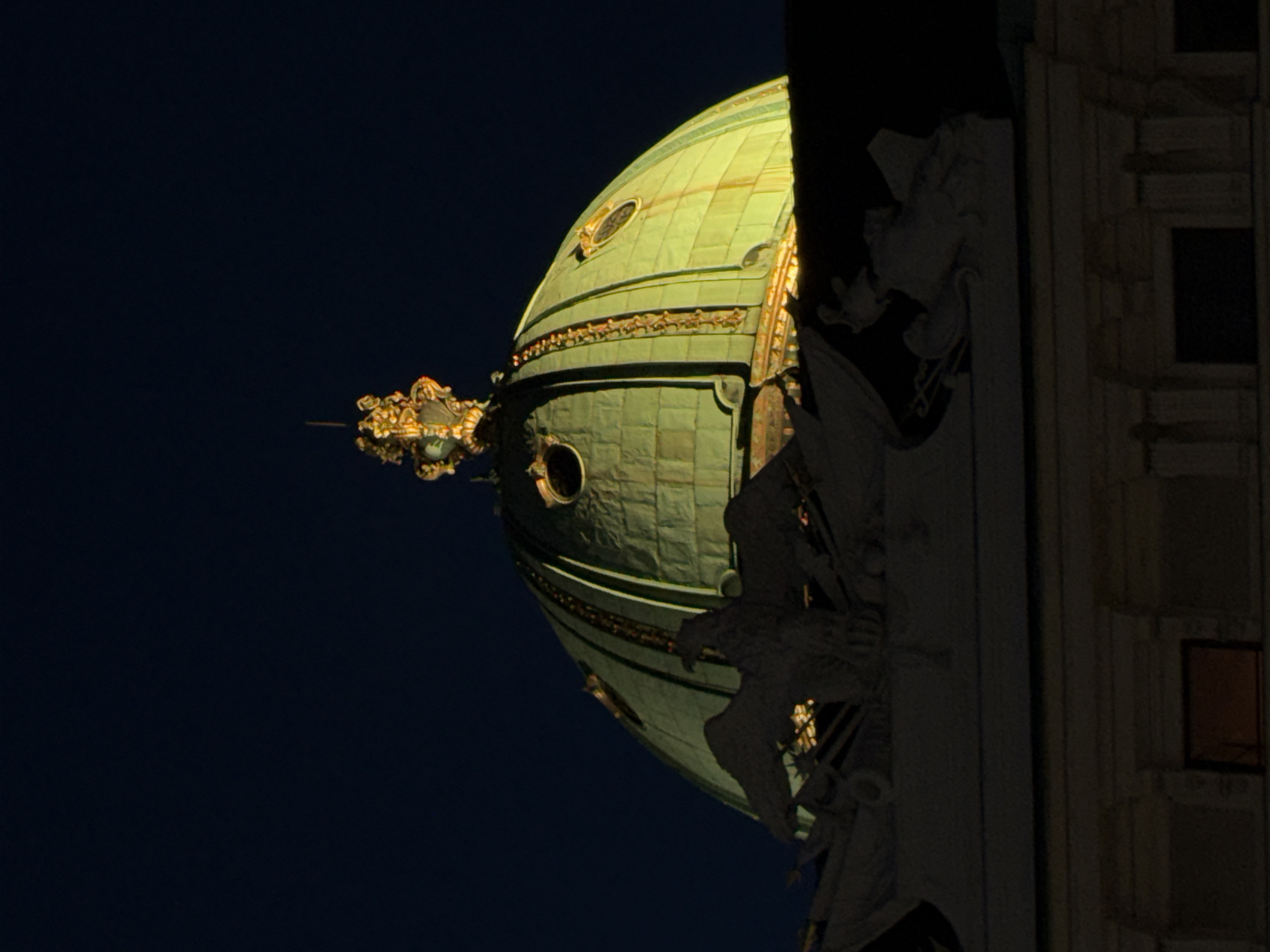 The image shows a close-up, low-angle shot of the dome of the Austrian National Library in Vienna, Austria. The dome is illuminated by artificial light, casting a green glow against the dark night sky. The dome is ornately decorated with gold accents and features several oval windows. In the foreground, silhouetted against the dome, is a stone sculpture of an eagle with outstretched wings. Below the dome and sculpture, the upper facade of a building is visible, with architectural details and darkened window openings. The scene appears to be set at night.