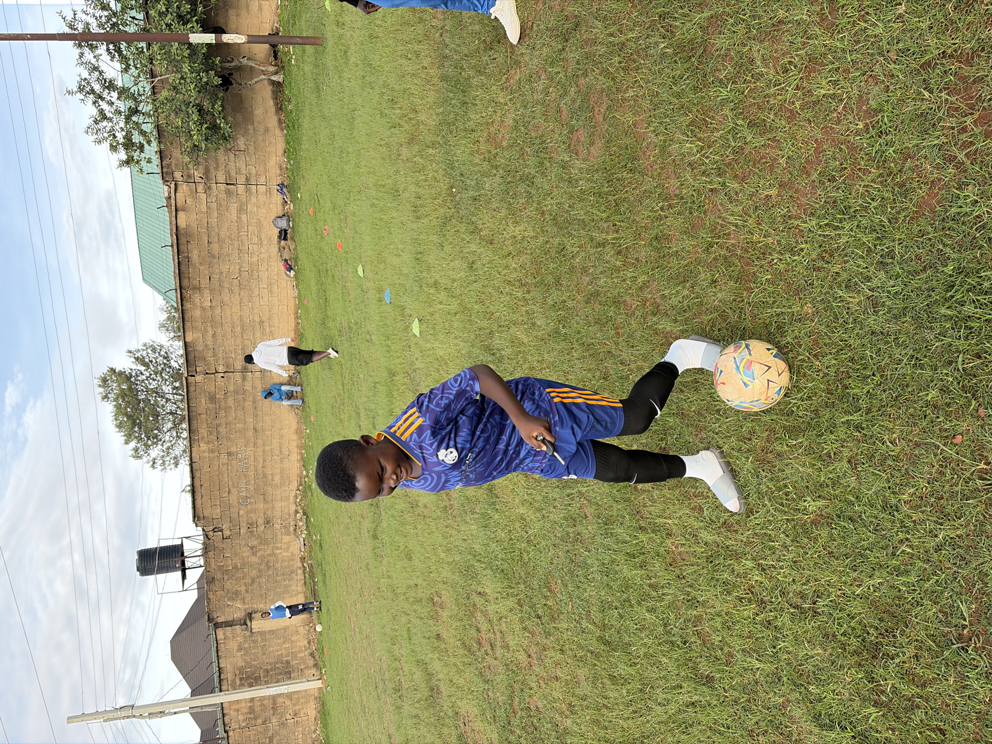 The image captures an outdoor scene on a grassy field in the city of Jos, Nigeria, under a partly cloudy sky. The primary subject, a young Black boy, estimated to be between 7-10 years old, is positioned in the foreground, slightly right of center. He wears a blue and gold Real Madrid soccer jersey, matching shorts, black compression leggings, white socks, and light-colored athletic footwear. He is actively engaging with a multi-colored soccer ball near his left foot, looking down with a smile, and holding a small, dark object in his right hand.
In the mid-ground, a weathered, light brown concrete block wall extends horizontally. The word "CAR PARK" is spray-painted in white on the wall to the left, and barbed wire is visible along its top. Behind the wall, structures include a building with a dark-tiled roof, a black water tank on a metal stand, and a building with a green roof, partially obscured by trees. Utility poles and power lines cross the upper frame.
Several individuals are present in the background: one person in a blue and white athletic uniform stands to the far left near a white soccer ball; another person in a light blue hooded sweatshirt sits against the wall in the center-right; and a person in a white long-sleeve shirt and black shorts walks towards the right. The lower legs of a fourth person in blue jeans and white shoes are visible at the far right edge. The grassy field is patchy, revealing reddish-brown soil in areas, and is scattered with small orange, green, and blue training cones. Miscellaneous items, including a backpack, are visible at the base of the wall.
