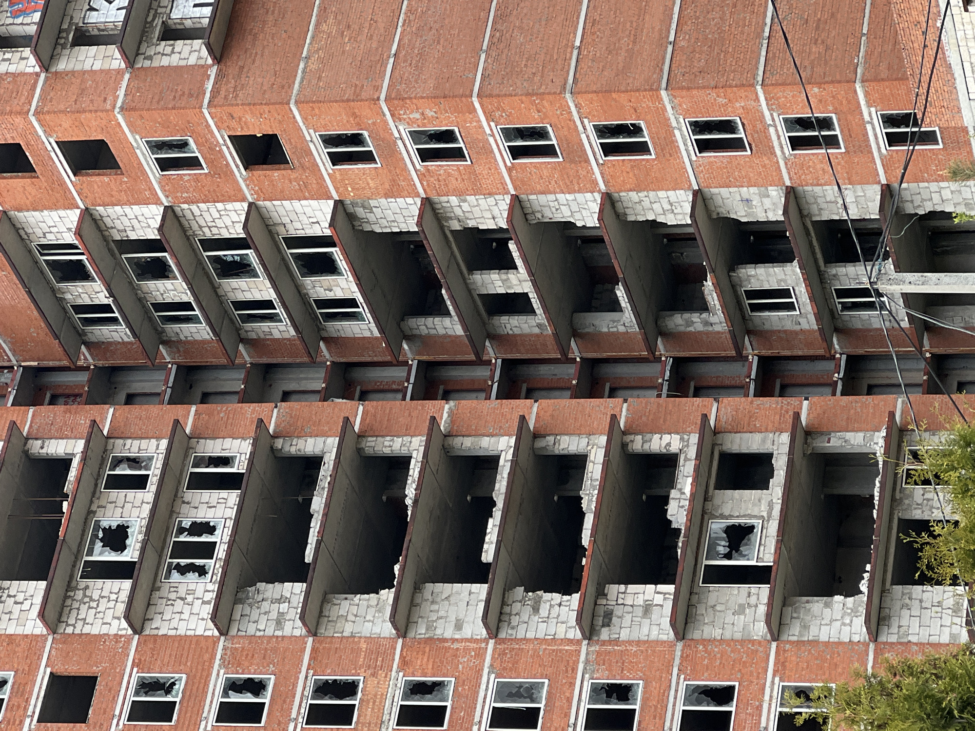 The image shows an exterior view of an abandoned and dilapidated building, likely a large apartment or office structure. The building is located in Perm, Russia. The facade is characterized by multiple rows of rectangular windows, many with shattered glass or missing panes. The building’s architecture includes deep, angled recesses between sections, creating a distinctive shadow pattern. The exterior walls appear to be constructed from a mix of red brick and light-colored concrete or block. There are some utility lines visible in the upper right portion of the image. There are some green leafy plants visible in the lower right corner of the image. There are signs of graffiti on the upper portion of the structure. The building's overall condition suggests long-term neglect and disuse.