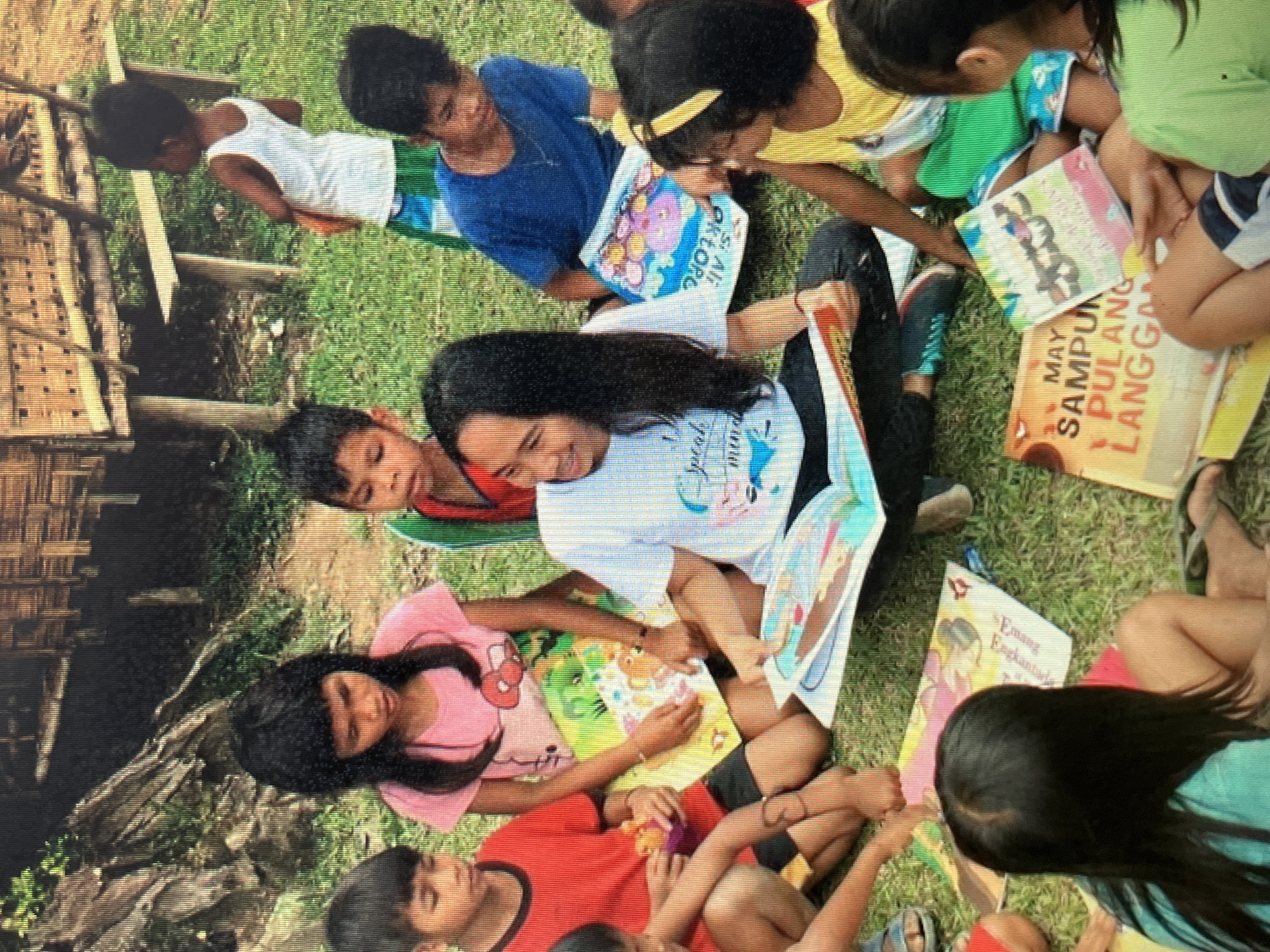 This image depicts an outdoor gathering of children and an adult in what appears to be a rural setting, possibly within the village of Sejahtera, Indonesia. The scene is set on a grassy area in front of a stilted wooden structure, likely a house or community building, with a dirt path leading away from it.
Several children, ranging in age from approximately five to twelve years old, are seated on the ground in a semi-circle. They are engaged in looking at books. Some are holding open colorful books, with one child pointing to the pages. The adult, a woman with long dark hair, is also seated and appears to be reading or interacting with the children from a book she is holding open.
The children are dressed in casual, brightly colored clothing. One child wears a red t-shirt, another a pink t-shirt with a character print, and a third a blue t-shirt. Another child is wearing a yellow and green outfit. An older child, possibly a teenager, is wearing a blue t-shirt. A young child in a white tank top and green shorts stands slightly apart.
The environment suggests a warm climate. The ground is covered in green grass, and the background shows a rocky embankment and dense foliage. The structure behind them is made of bamboo and wood, characteristic of traditional Indonesian architecture. The overall atmosphere is one of a community learning or storytelling session.
