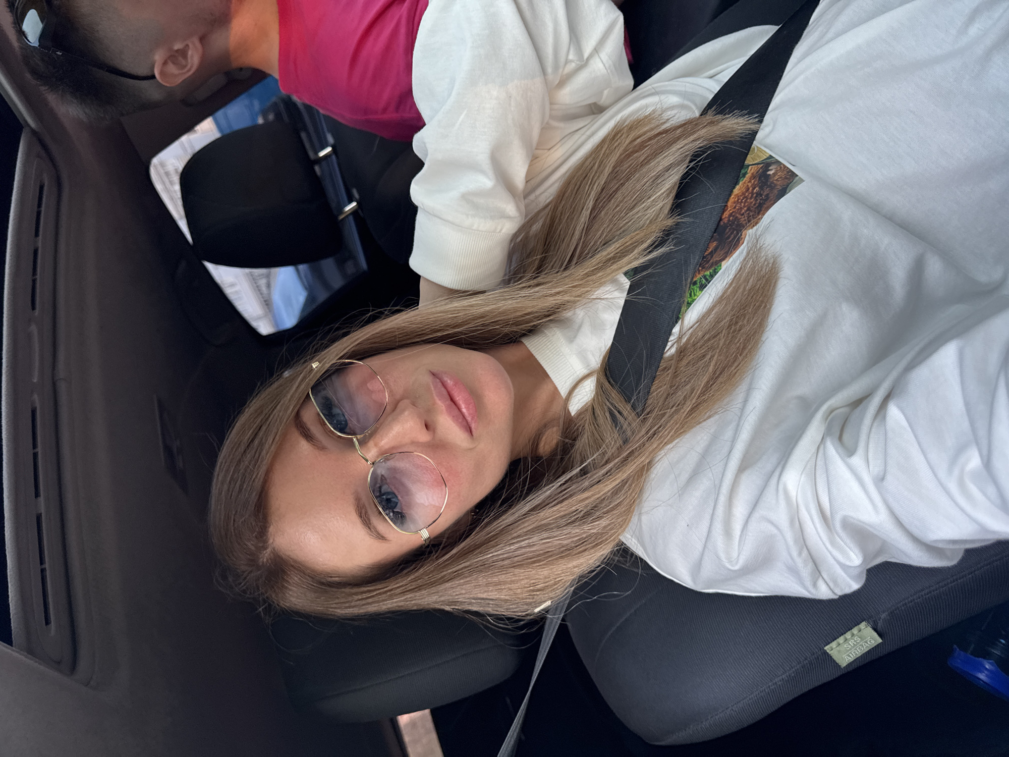 A woman with blonde hair and glasses is taking a selfie in the passenger seat of a car. She is wearing a white sweatshirt. A man in a pink shirt is visible in the background, also in the car. The car appears to be moving, as indicated by the background blur. The image was taken on a sunny day, with natural light illuminating the scene. The woman's hair is styled loosely around her shoulders, and she is looking directly at the camera with a slight smile. The car's interior is dark, with the exception of the light reflecting off the woman's glasses. The seatbelt is visible across her chest. The specific location in the city of Chaikovsky, Russia, cannot be determined from the visual information provided in the image.