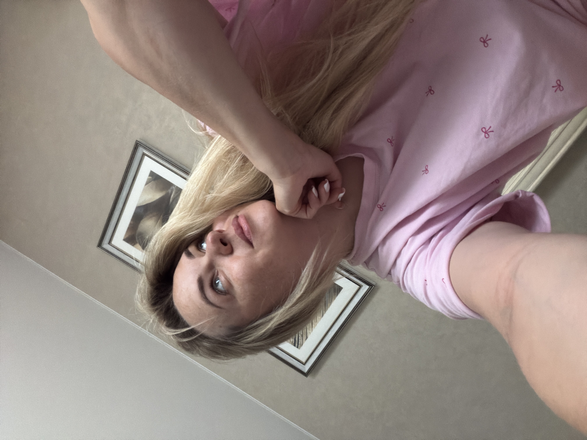 The image displays a head-and-shoulders portrait of a person with long blonde hair, taken from a low-angle perspective and rotated clockwise, causing the subject and background elements to appear on an angle. The person is looking directly upwards (towards the top left of the rotated frame) with blue or grey eyes, and has a neutral to slightly contemplative expression. Their left hand is raised to their chin/jawline, displaying fingernails painted white. They are wearing a light pink top adorned with small, repeating darker pink bow patterns. The skin tone is fair.
In the background, behind and above the person's head (in the rotated view), there are two framed pictures hanging on a light, neutral-colored wall, likely beige or light grey. The frames appear to be silver or light-colored, and one of the visible images within a frame seems to depict a plant or an abstract pattern. The setting appears to be indoors, possibly a room in a home. No specific activity beyond posing for the photograph is evident. The time of day cannot be definitively determined, but the lighting suggests an indoor environment, possibly daytime. There is no visible text. The location context provided is Votkinsk, Russia.