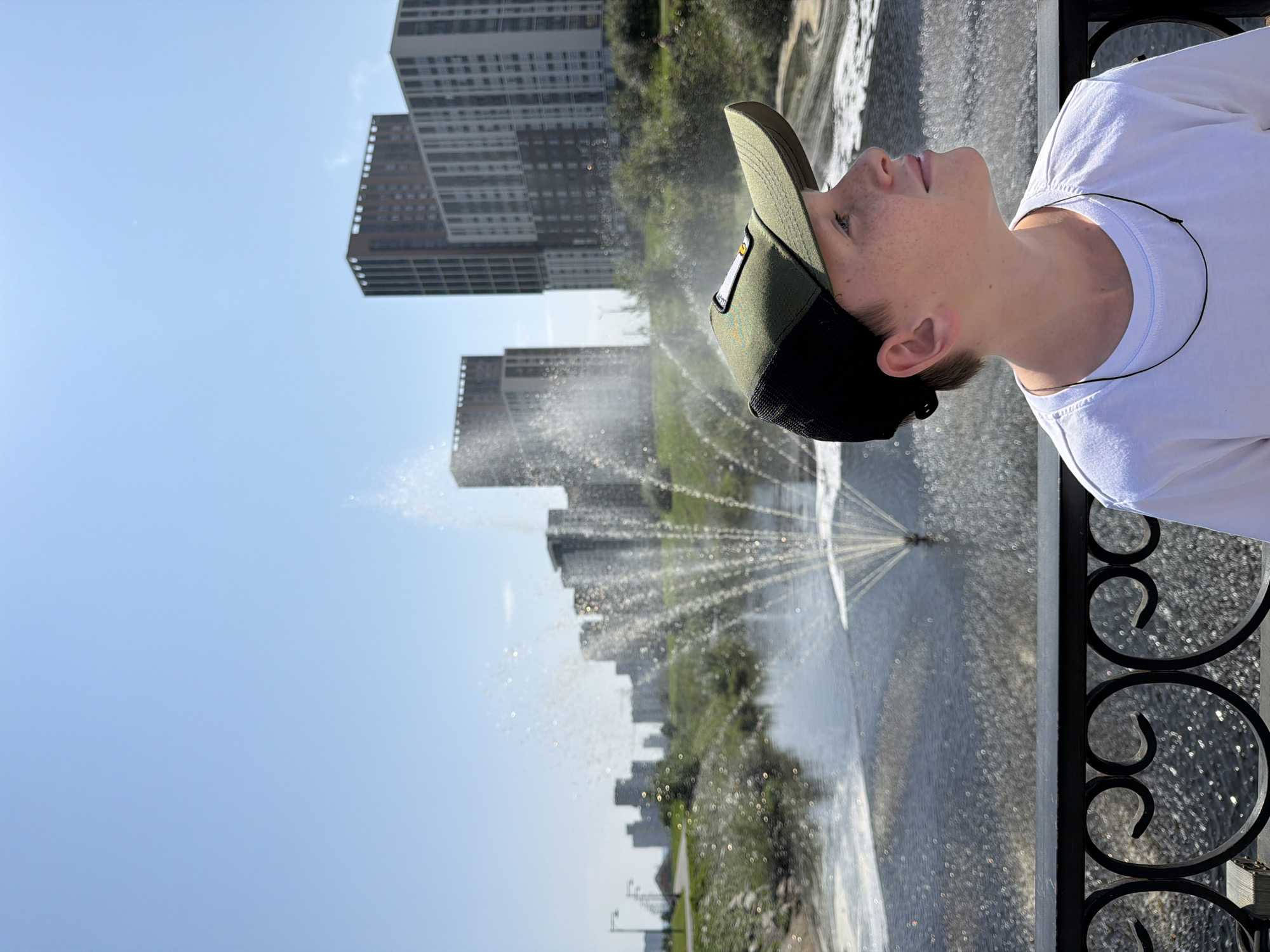 A young Caucasian boy wearing a green and black baseball cap and a white t-shirt stands on a bridge in the city of Yekaterinburg, Russia. He is looking to his right, away from the camera. In the background, a large fountain sprays water into a body of water. Behind the fountain are several modern high-rise apartment buildings and a clear blue sky. The bridge railing is made of black metal with decorative scrollwork. The overall scene depicts an outdoor urban environment on a sunny day.