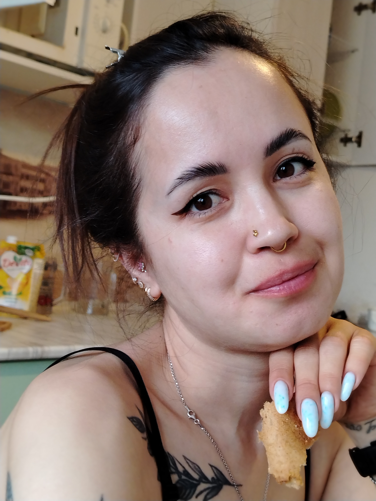 A close-up selfie features a fair-skinned woman with dark hair pulled back into a loose bun, secured with a hair clip. She has dark, defined eyebrows, brown eyes with eyeliner, and a small gold nose ring. She is smiling gently. Her manicured nails are long and oval-shaped, with a light blue, cloud-like pattern. She is holding a piece of bread in her left hand, which is propped under her chin. She is wearing a black tank top and a silver necklace. Tattoos are visible on her chest and arm.
The background is a kitchen setting, with white cabinets and a partial view of what appears to be a brick accent wall. A yellow package is visible on the countertop behind her.
The location is not mentioned in the picture, and hence it is impossible to tell if the scene is from the city of Noviy, Russia.