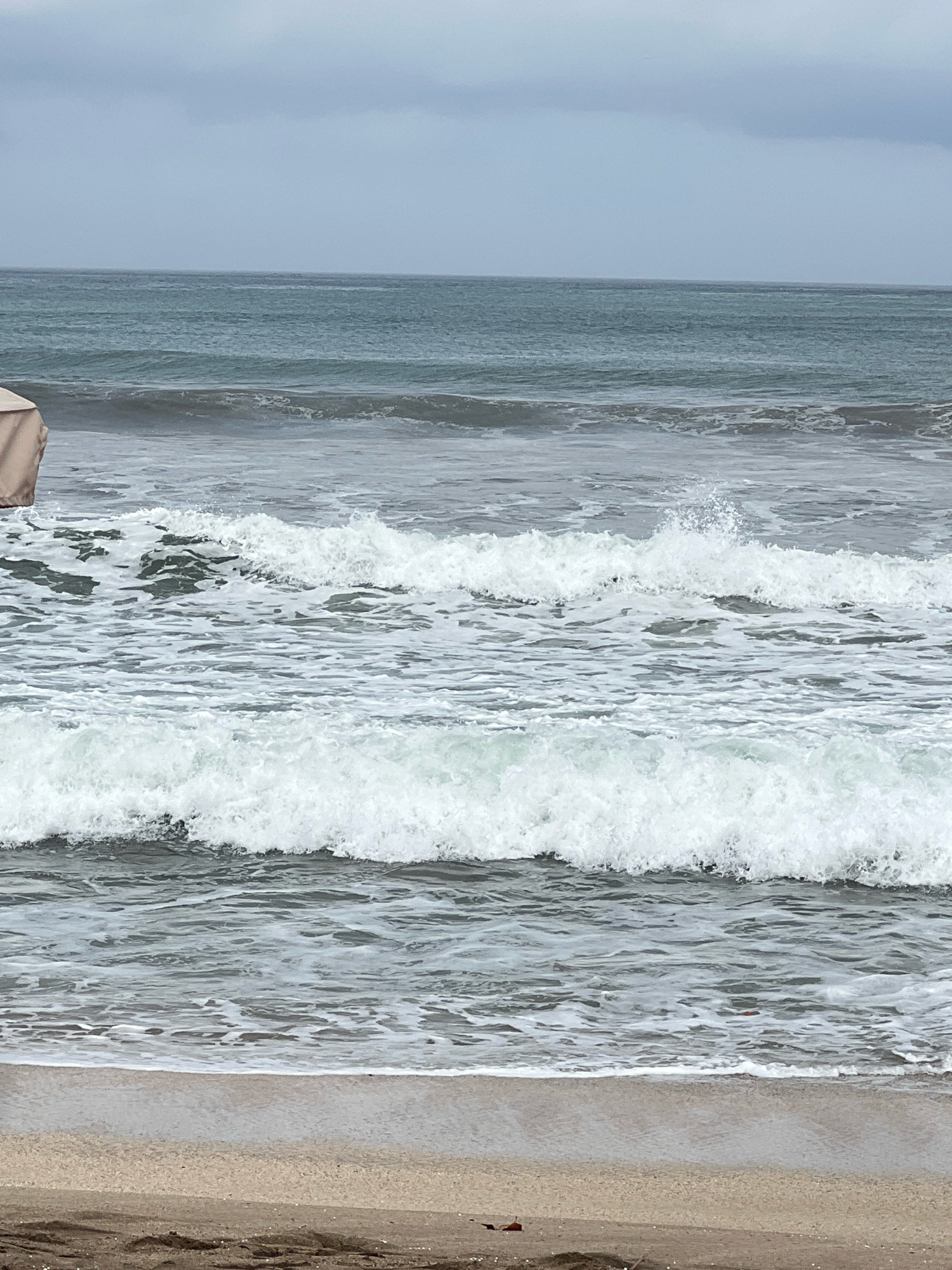 A wide shot captures the turbulent ocean waters at Kuta beach in Indonesia under an overcast sky. Waves crash and foam rolls ashore onto the sandy beach. In the upper left corner, a sliver of a light-colored beach umbrella is visible. The horizon is a faint line where the grey sky meets the dark ocean.