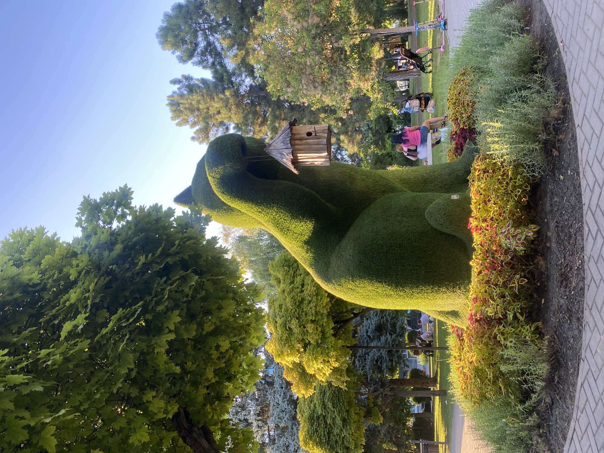 A large, topiary sculpture of a cat holding a wooden birdhouse in its paw is the central subject of this image, set in a park-like environment. The cat is a vibrant green, meticulously trimmed, and sits on a bed of colorful flowers, primarily coleus with red and yellow foliage, and some green, shrub-like plants. Behind the sculpture, a paved walkway is visible, leading into a grassy area with several trees. In the background, two individuals are seated on a bench, their backs to the camera, with a stroller nearby. Further in the distance, other park-goers, including children on scooters, can be seen enjoying the outdoors. The sky is clear and blue, with sunlight illuminating the scene, casting shadows and highlighting the textures of the foliage. This scene is located in Chișinău, Moldova.