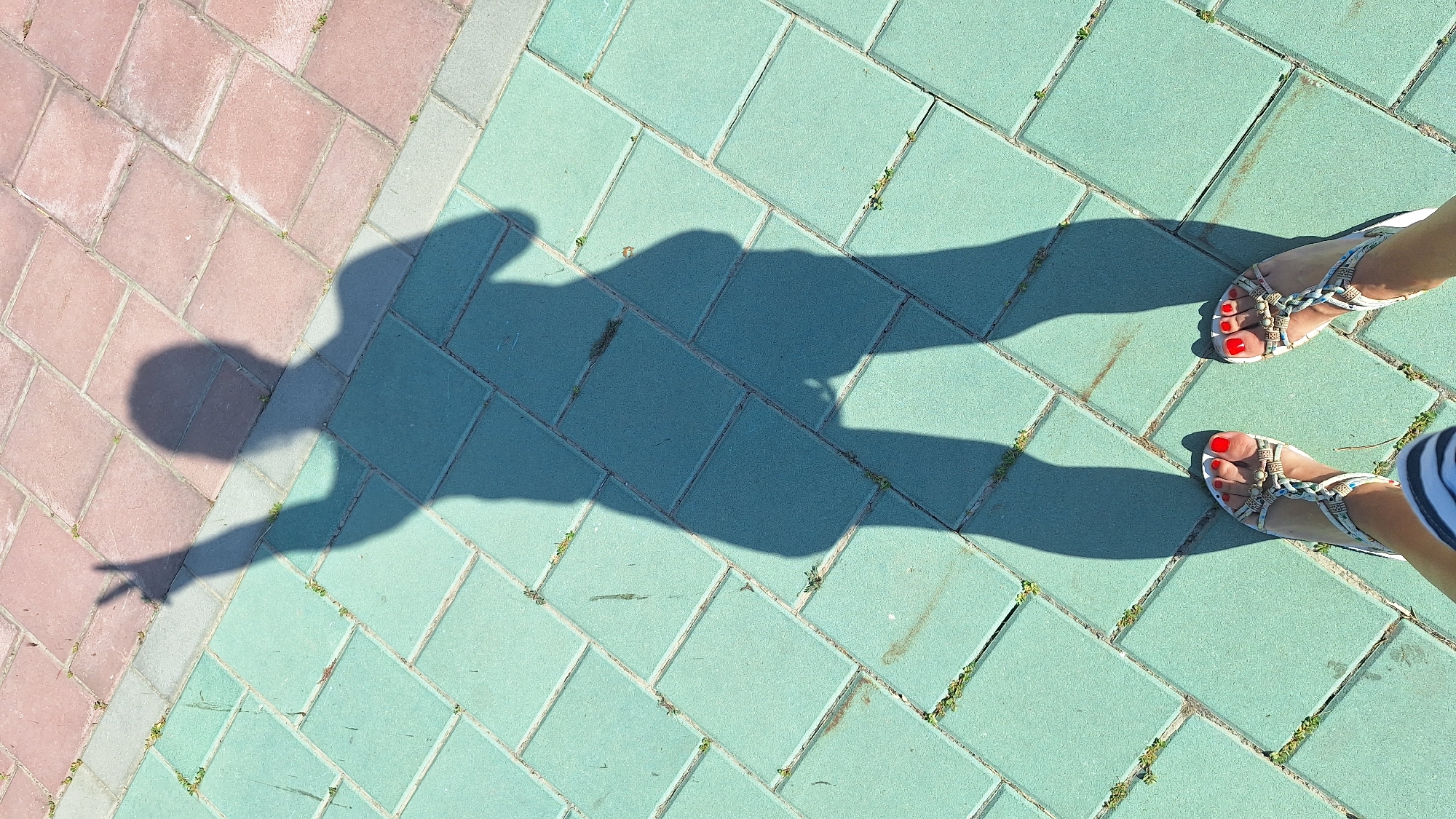 The image captures a shadow of a person with their right arm raised, index and middle fingers extended in a peace sign, against a backdrop of interlocking pavement stones. The paving stones are arranged in a diagonal pattern, with alternating sections of reddish-brown and teal-green. Small patches of grass are visible growing in the gaps between the stones. The bottom of the frame shows a pair of feet wearing sandals with a white and blue patterned design, and red-painted toenails. The scene is lit by sunlight, casting a distinct shadow. The setting is likely an outdoor public space such as a walkway or plaza within the city of Sombor, Serbia.