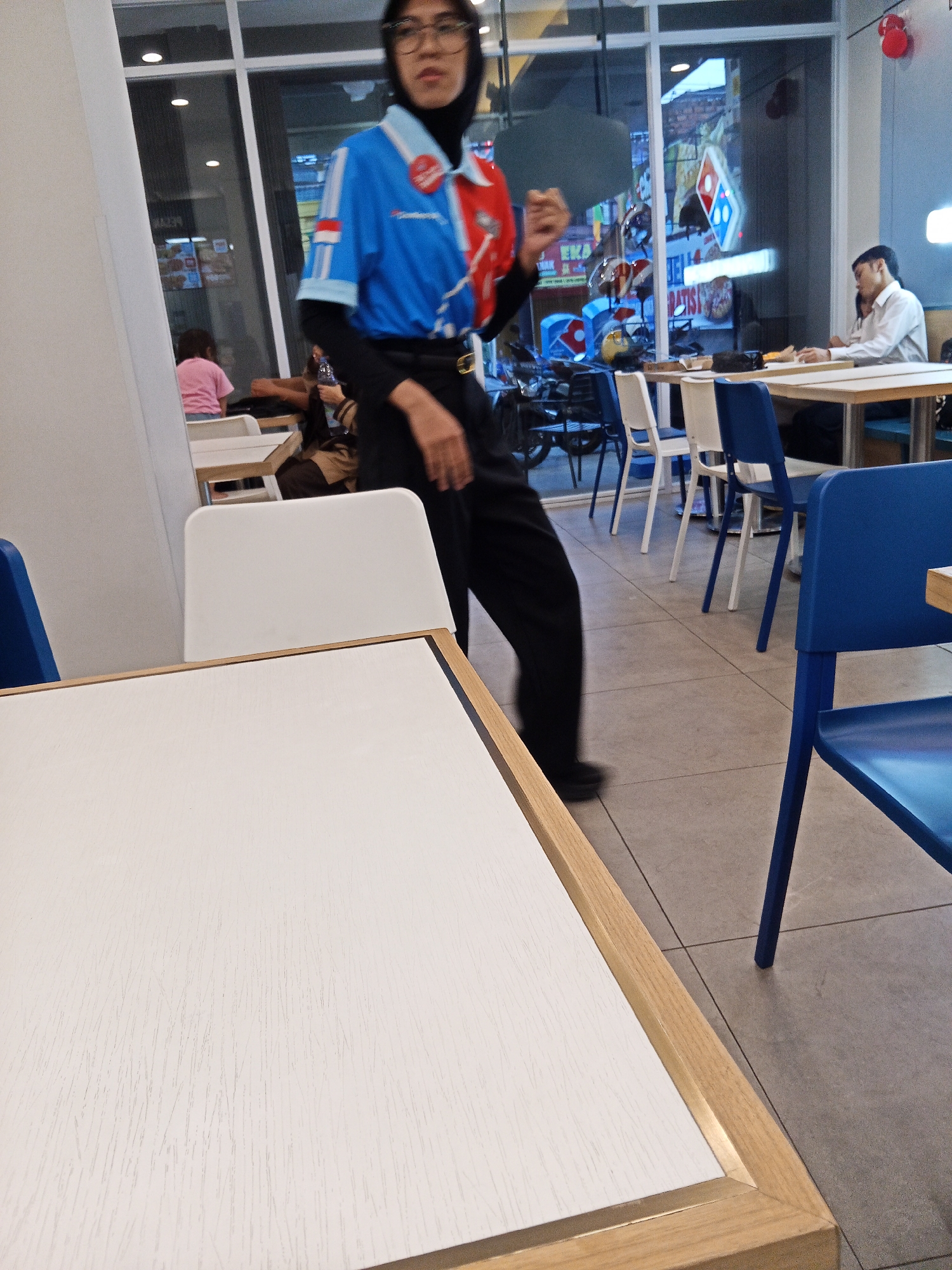 The image depicts the interior of a Domino's Pizza restaurant in Lubuklinggau, Indonesia. A person wearing a black hijab, glasses, and a blue and red Domino's uniform is walking across the tiled floor. They are wearing black pants and a belt. In the background, several tables and chairs are visible. To the right, a man in a white shirt is seated at a table, talking on his phone. Through the large glass windows in the background, the exterior of the restaurant can be seen, including other Domino's signage, motorcycles, and other businesses. The lighting suggests it is daytime.