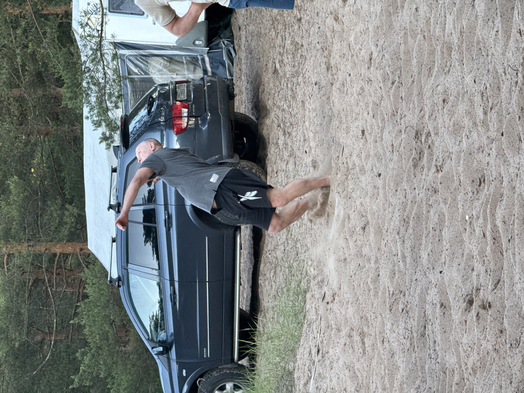 A man is mid-throw while playing a game in the sand at a campsite. He is wearing a grey t-shirt and black shorts. The man's body is arched backwards, and his right arm is extended upwards, having just released an object. Sand is kicked up around his feet, suggesting vigorous movement. Behind him, a dark grey SUV is parked on the sand, with a portion of a white caravan visible to the right. Trees form a backdrop to the scene. The image appears to be taken in Ozerki (Krasnodolinskaya), Russia, based on the provided context.