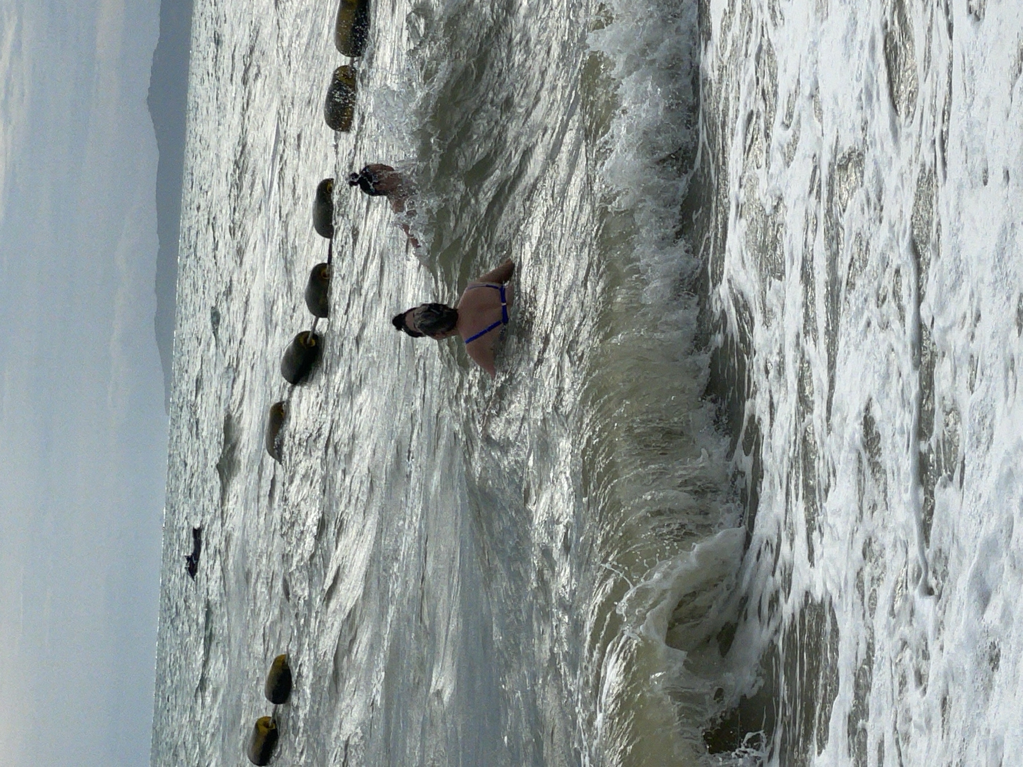 The image depicts a choppy sea with moderate waves near the shore, likely in Pattaya City, Thailand. A line of buoys is visible in the mid-ground, extending horizontally across the frame. In the foreground, a wave is breaking, creating white foam. Two individuals are in the water. One person, facing away from the camera, is wearing a blue bikini top and appears to be swimming. Another person, also facing away, is further back in the water, with water splashing around them. In the far background, a silhouette of landmass, possibly mountains, is visible against a cloudy sky. A small dark object, potentially a boat or jet ski, is seen in the distance on the water.