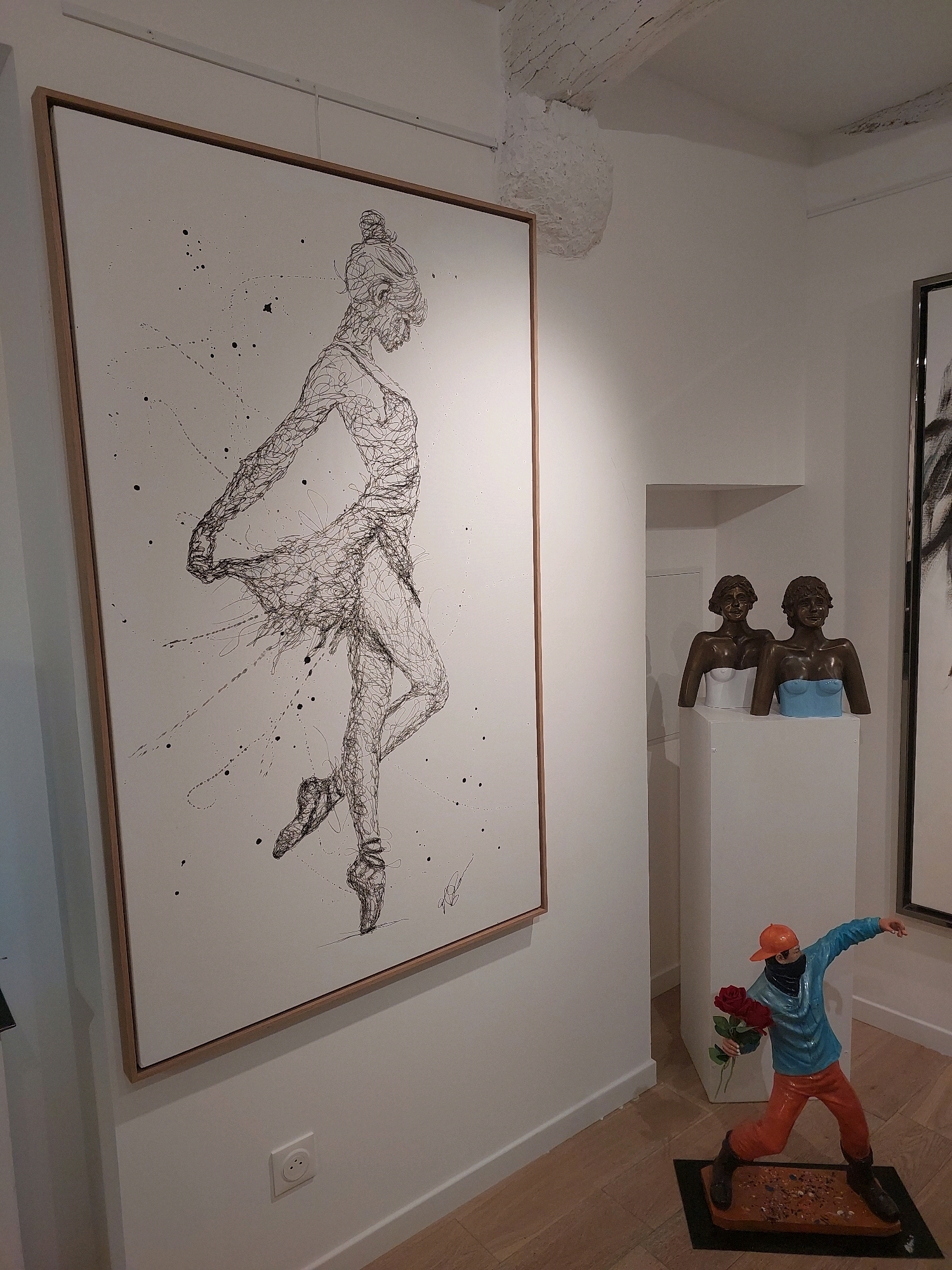 The image depicts an interior scene, likely an art gallery, in the city of Saint-Paul-de-Vence, France. Dominating the left side of the frame is a large, framed artwork of a ballerina drawn with thin, black lines on a white background. The drawing is dynamic, with the ballerina in a pose of movement, her form fluid and somewhat abstract, with splashes of black ink scattered around her.
To the right of the painting, there is a white pedestal displaying two bronze busts of women. The bust on the left has a white top, while the one on the right wears a light blue top. Both women have dark, curly hair and appear to be smiling.
In the foreground on the right, a colorful figurine of a boy wearing an orange cap, a blue jacket, and orange pants is positioned on the floor. He is shown in a throwing stance, holding a bouquet of three red roses in his left hand. He stands on a dark base adorned with small, irregular objects. The floor is made of light-colored wooden planks, and the walls are painted white. The ceiling features exposed wooden beams and some white, textured material hanging from above. Another framed artwork, partially visible on the far right, suggests more art is displayed in the space.