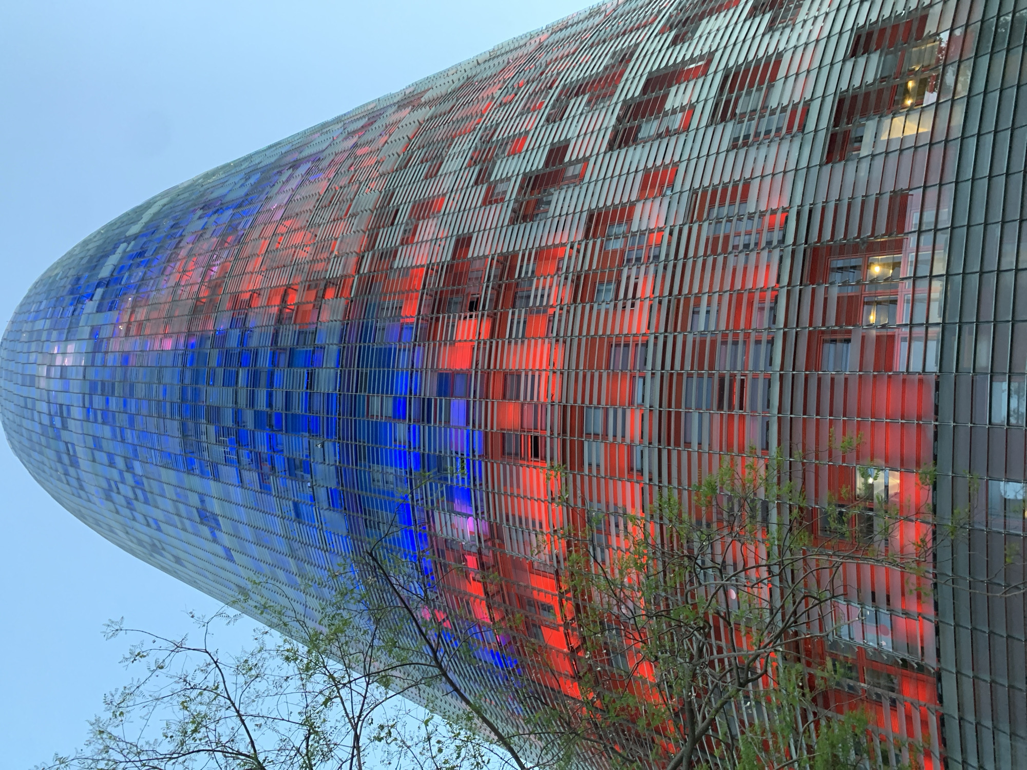 The image shows the Agbar Tower in Barcelona, Spain. The building is a cylindrical tower with a domed top, and its facade is covered in a mosaic of colored panels, primarily blue on one side and red on the other. Some greenery is visible in the foreground, obscuring parts of the building's lower levels. The sky is a pale blue.