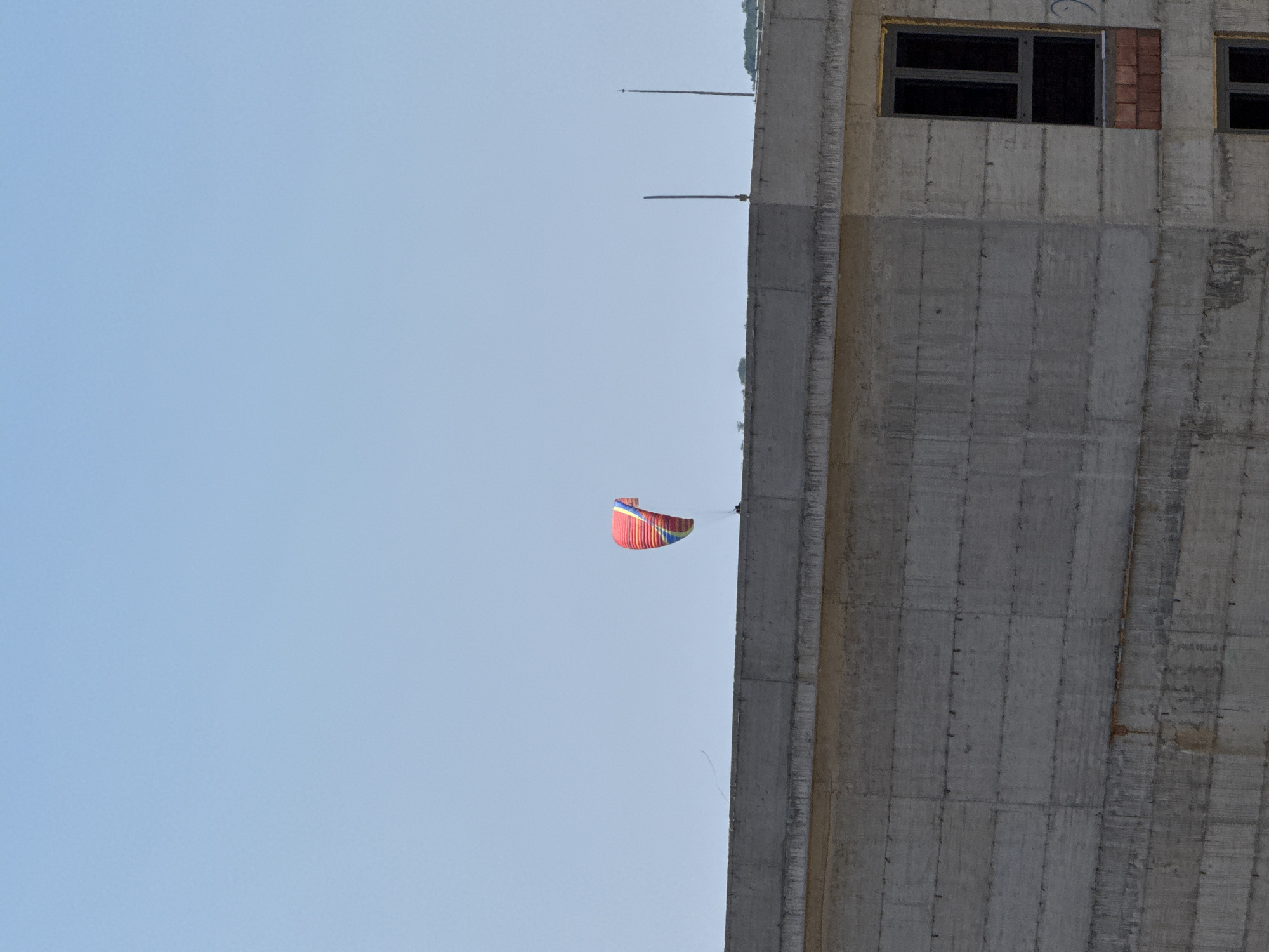 A person is visible in the sky, in the process of kiteboarding. The kite is red, blue, and yellow. The person is elevated above a concrete structure. The concrete structure appears to be under construction, with visible rebar protruding from the top. The sky is clear and light blue. The background suggests a hilly landscape, with trees visible on the horizon. The scene is likely located in Boreti, Montenegro.