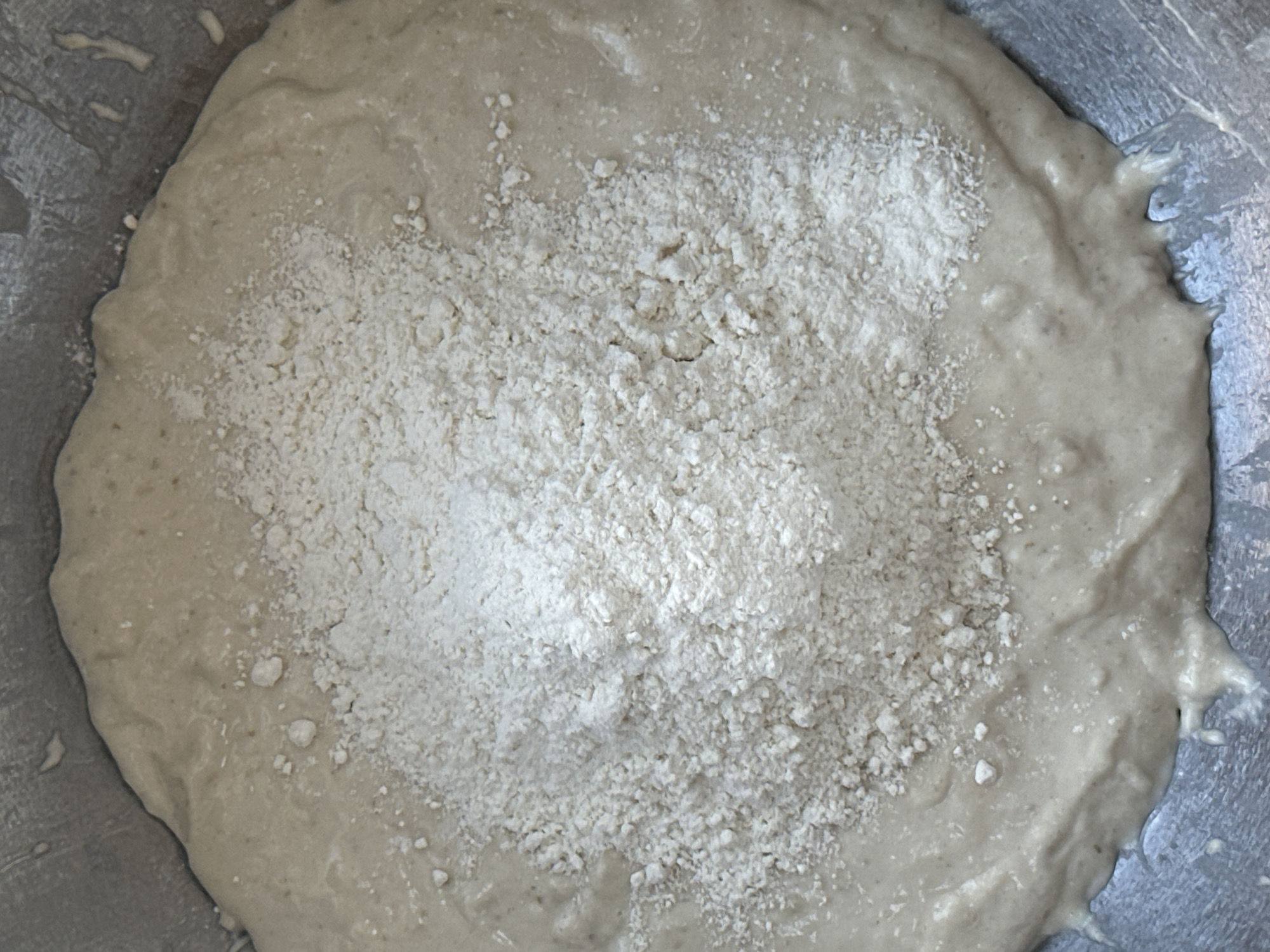 The image shows a close-up, top-down view of what appears to be dough in a metal bowl. The dough is light beige in color and covers most of the bowl's surface. A large pile of white flour sits in the center of the dough. The surrounding environment is not discernible, and there is no information available to determine the location of the image within Wealden, United Kingdom, or any details about people or interactions. The primary objects are the dough, the flour, and the bowl.