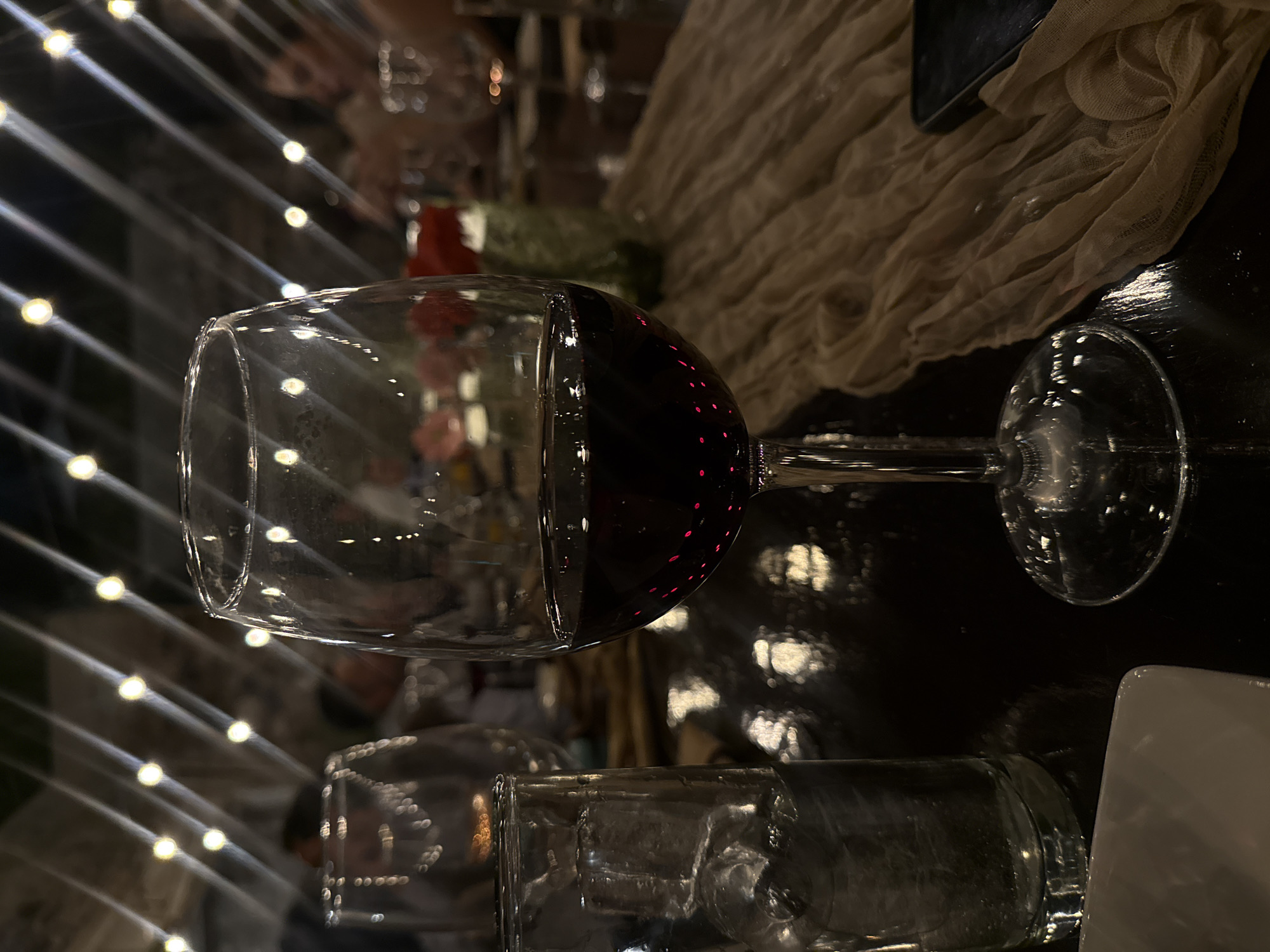 A red wine glass, partially filled with red wine, sits on a dark, reflective surface in Santiago de Querétaro, Mexico. Next to it is a water glass and a white plate. A beige fabric is draped in the background. The scene is illuminated by string lights, creating a warm ambiance. Other glasses and tableware are visible in the blurred background.