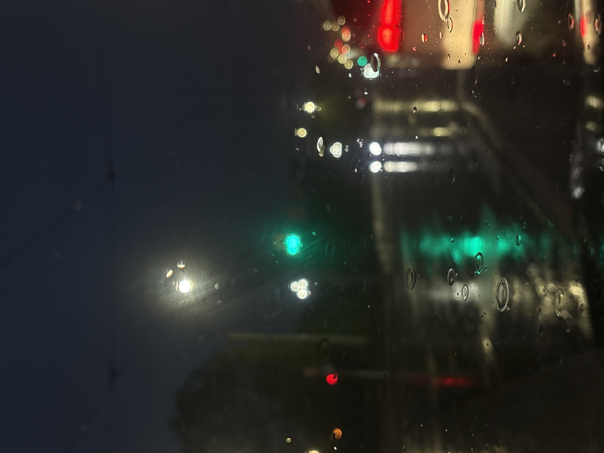 The image depicts a view through a rain-streaked window at night, likely from inside a vehicle. Out of focus, there are various light sources, including traffic lights (red, green, and possibly yellow), streetlights, and vehicle headlights/taillights. The environment appears to be urban, possibly a street or intersection. The city is reported to be Minsk, Belarus, but nothing definitively in the image can prove this. The raindrops on the window obscure and distort the view. No people or direct interactions are visible.