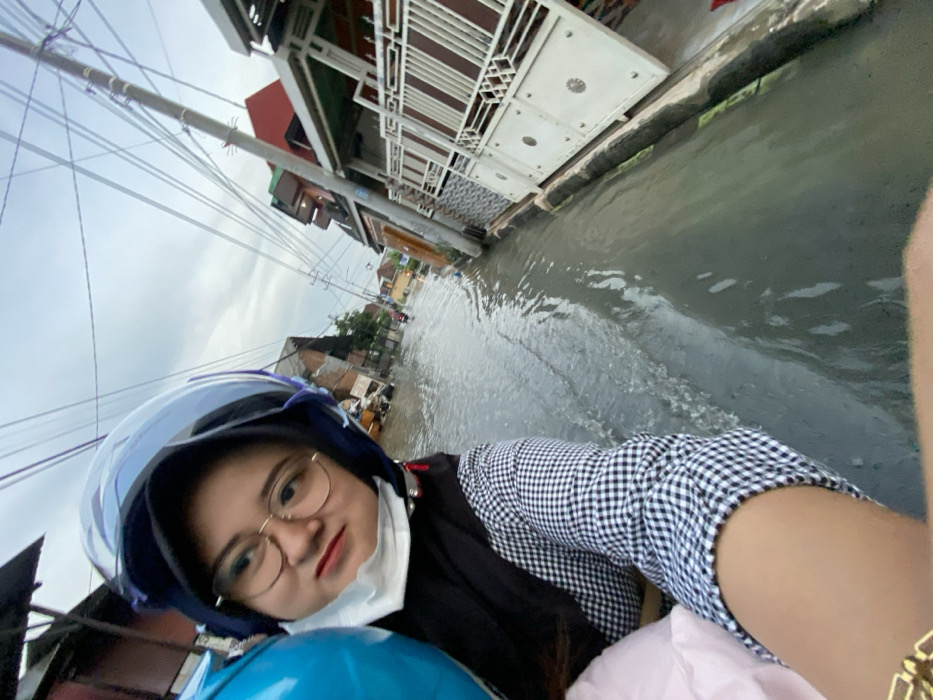 This image captures a flooded street scene in Jambangan, Indonesia, from the perspective of a person on a motorbike. In the foreground, a female individual wears a dark blue motorcycle helmet with a clear visor, round gold-framed glasses, a white surgical mask pulled down to her chin, a black head covering, a black vest, and a white and black gingham long-sleeved shirt. Her gaze is directed slightly to the right of the camera. A blue, possibly inflatable, object is partially visible to her left, while her right forearm, adorned with a gold-colored bracelet, is visible in the lower right.
The street is inundated with murky grey floodwater, appearing several inches deep, with visible ripples and displacement indicating motion. Residential buildings line both sides of the submerged road. Structures on the left exhibit varied construction, including exposed brick and corrugated materials. On the right, more established residential buildings are visible, featuring multiple stories, white ornate metal gates, and colorful exterior elements. A series of utility poles with a dense network of overhead cables stretch across the frame. The sky above is overcast. In the background, faint figures or vehicles are discernible navigating the flooded thoroughfare.