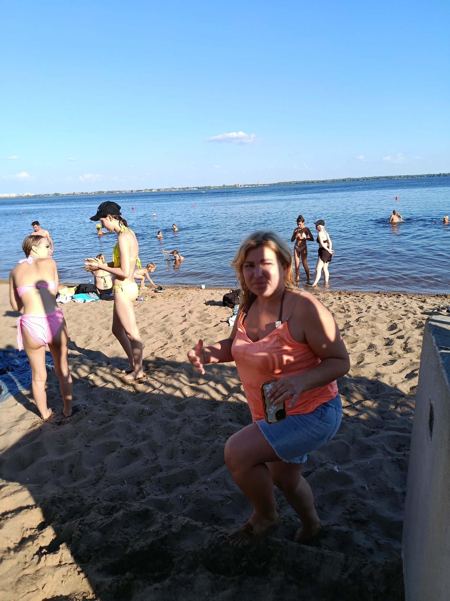 A group of people are at a beach on a sunny day. The sky is blue with some scattered clouds. The water is calm and blue. There are several people in the water, some swimming and some standing. On the beach, there are a few people sitting and standing. The sand is light brown and appears to be somewhat wet near the water. In the foreground, a woman in an orange tank top and denim shorts is looking towards the camera and holding a cell phone. To her left, two young women in bikinis are standing and talking. In the background, more people are visible in the water and on the shore. The overall scene depicts a relaxed summer day at the beach.