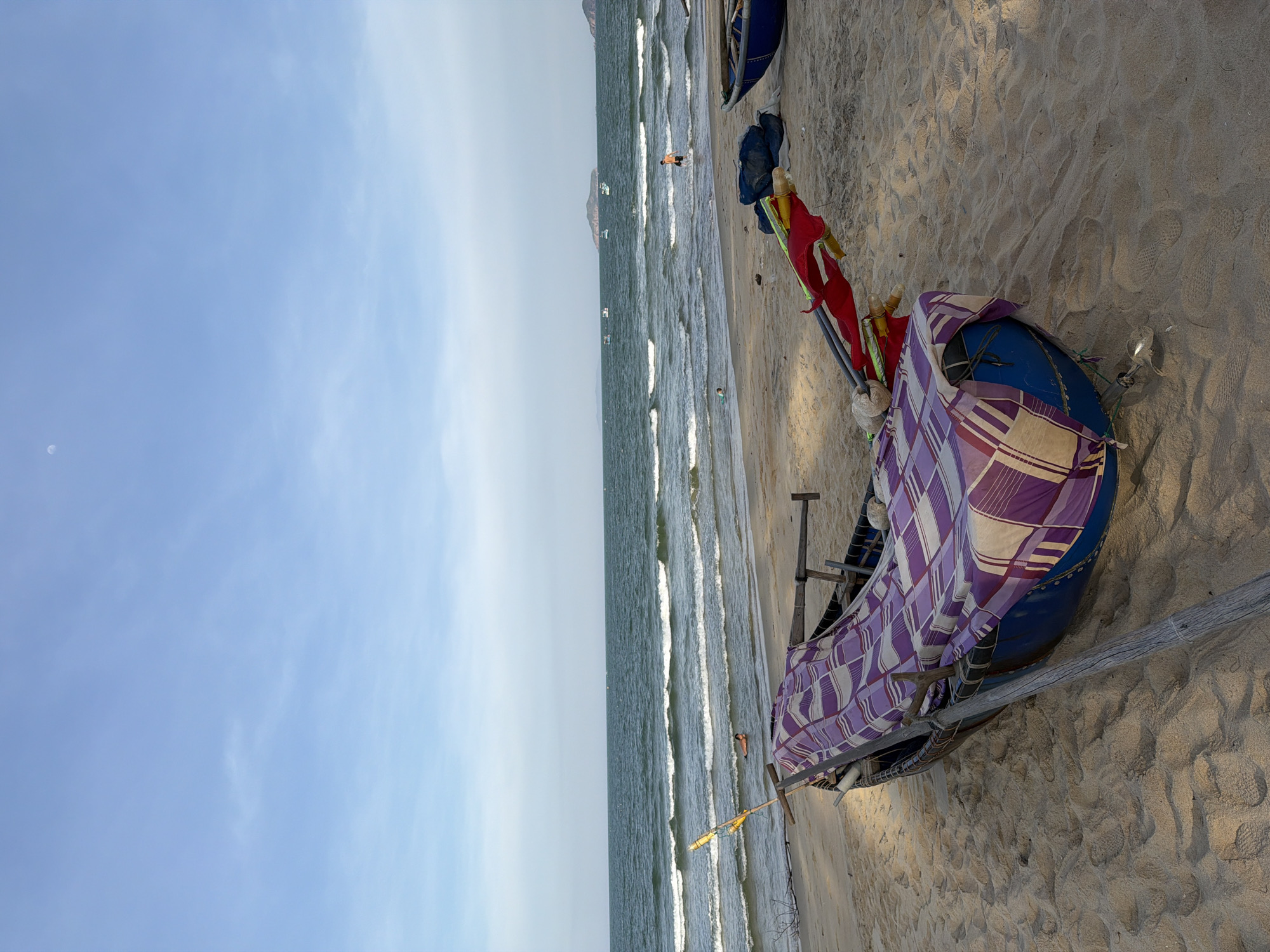This media file depicts a serene beach scene in South Quy Nhon Ward, Vietnam. The image is oriented vertically, showcasing a vast expanse of clear blue sky on the left and the ocean and sandy shore on the right. A crescent moon is faintly visible in the sky.
The ocean is calm with gentle waves lapping the shore, suggesting a peaceful time of day, likely late afternoon or early morning given the soft lighting. A few small boats are scattered in the water, and one person is standing near the water's edge in the distance, while another appears to be swimming.
In the foreground, a fishing boat rests on the sandy beach, partially covered with a purple and white checkered cloth. Two poles with red flags are propped up beside it, possibly for signaling or as part of the fishing equipment. The sand is textured with footprints and the gentle ripples left by the receding tide. The overall atmosphere is tranquil and idyllic, highlighting the natural beauty of the Vietnamese coast. There is no visible text in the image.