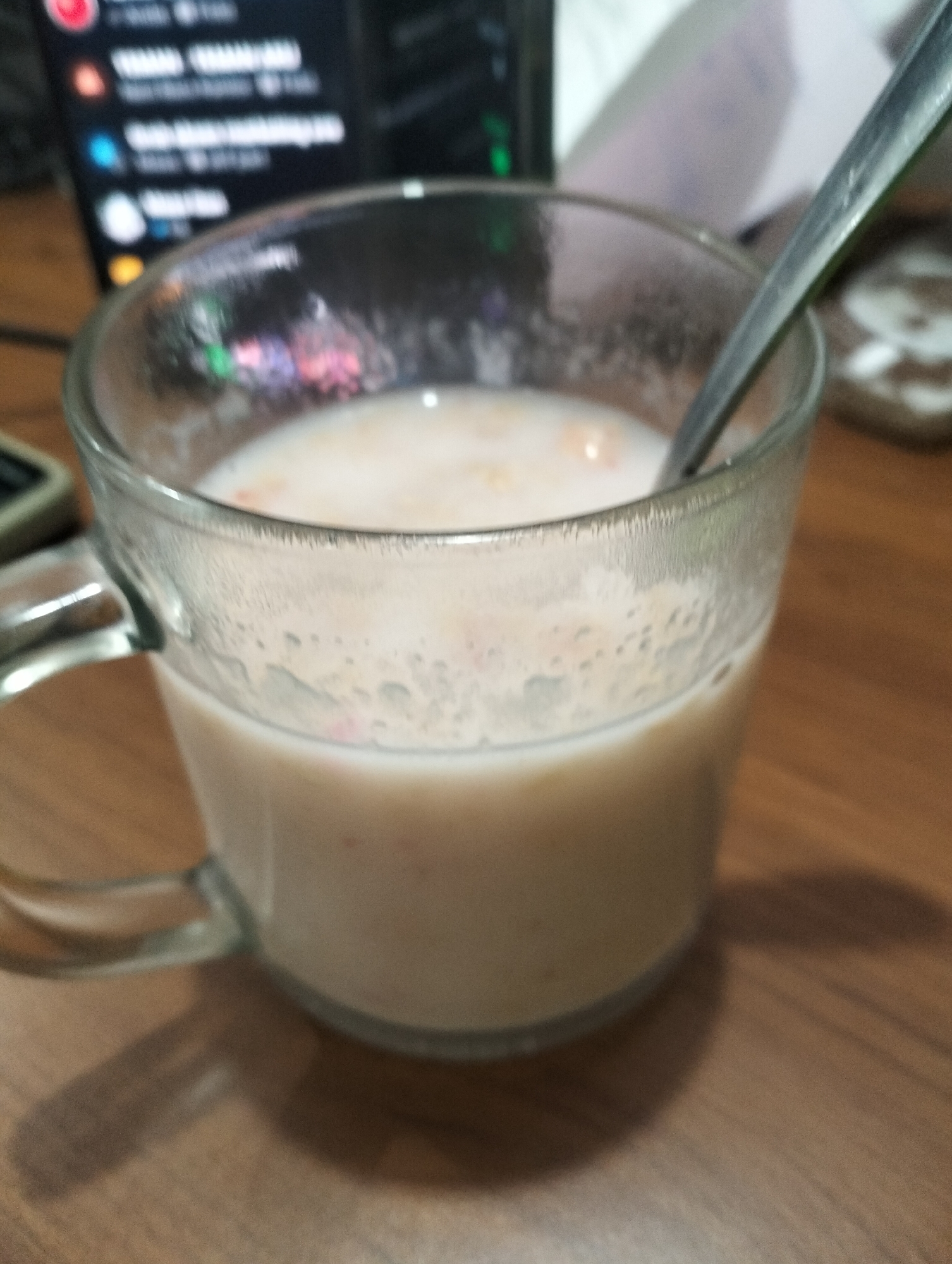 A close-up, medium-shot photograph shows a clear glass mug filled with a milky beverage, likely a cereal drink or smoothie, with some small pieces of fruit or cereal visible within. A metal spoon is partially submerged in the drink, resting against the rim of the mug. The mug has a handle and is placed on a wooden surface. In the blurred background, a dark screen with glowing white and colored text is visible, suggesting a computer or monitor. To the right of the mug, a portion of a white plate with a brown pattern is visible. The lighting suggests an indoor environment, possibly a desk or table setting. The photograph does not provide enough information to determine the specific location within Wedoro, Indonesia.