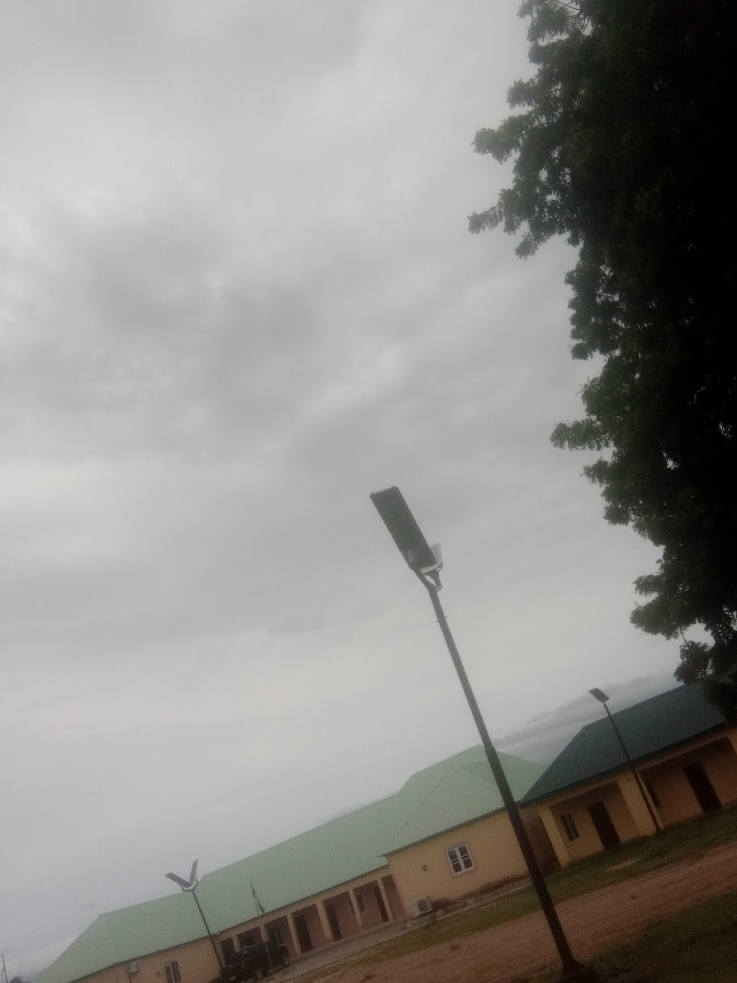 The image depicts an outdoor scene under a cloudy sky, likely in Jalingo, Nigeria. A dark-leaved tree dominates the upper right portion of the frame, its branches casting shadows. In the foreground and midground, there are buildings with light yellow walls and green roofs. These appear to be single-story structures, possibly an institutional or residential complex. A street lamp with a dark, rectangular fixture stands prominently in the center, angled upwards. Another street lamp, smaller and with a V-shaped top, is visible on the left, in front of the buildings. The ground is a mix of dirt and sparse grass. A portion of a vehicle is visible between the buildings. The weather appears overcast, suggesting it might be early morning or late afternoon, or simply a cloudy day. There is no visible text in the image. The overall impression is one of a somewhat subdued, possibly humid, environment.