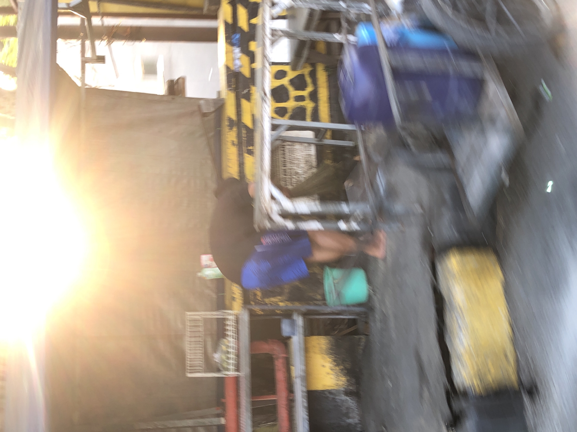 The image depicts an outdoor scene under bright sunlight, likely in a market or commercial area. A person, wearing a dark shirt and blue shorts, is bent over, possibly engaged in some activity. In the foreground, there is a wheeled cart with large blue containers. The background shows a structure with a dark tarp, yellow and black striped barriers, and shelves holding various items, including a white rack with what appears to be produce. A green bucket is visible near the person's feet. The environment appears somewhat gritty, with a concrete or asphalt ground. The scene is located in the city of Ngaban, Indonesia.