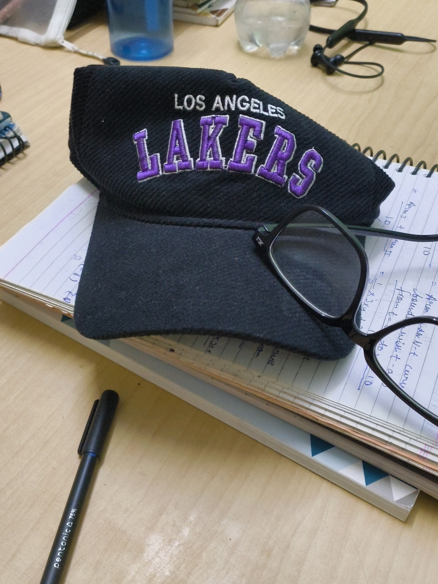 A black Los Angeles Lakers baseball cap is placed on top of a stack of notebooks on a wooden desk. The cap has the words "LOS ANGELES" and "LAKERS" embroidered in purple. Next to the cap, on the notebooks, are a pair of black-rimmed glasses and a black pen. The pen is labeled "pentonic 75%". In the background, there is a blue water bottle and a pair of black wireless earbuds. The image appears to be taken indoors, likely in a home or office setting. There is no indication of the city of Bulandshahr, India, or any other specific location.