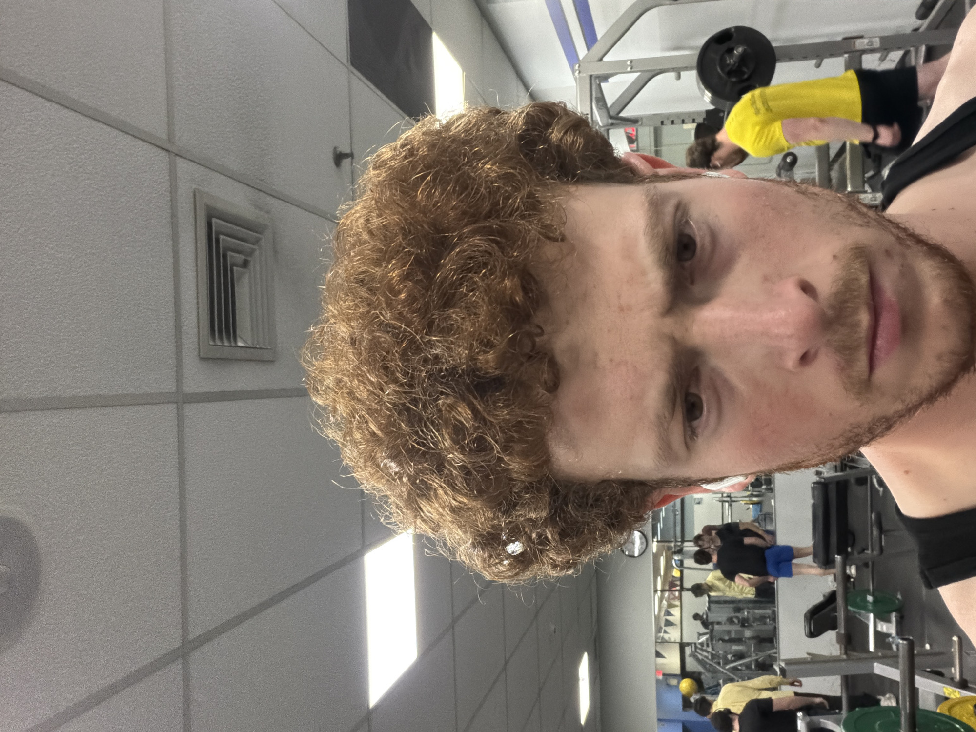 The image is a selfie taken by a person in a gym setting. The individual has light skin, brown hair, and a beard. They are wearing a black tank top. The gym has white tiled walls with recessed lighting and air vents. Other people can be seen in the background, some using weightlifting equipment. A barbell with weights is visible on a rack. The location is likely a gym somewhere in the city of New York, United States of America.