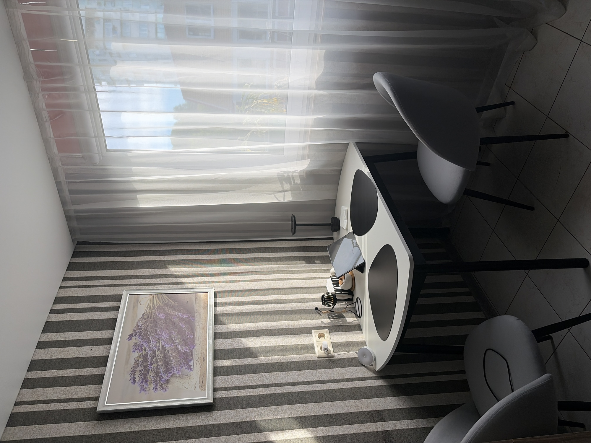 This image presents an interior view of a compact dining or breakfast nook, located within a residential setting in Sochi, Russia. The room features a prominent wall covered with vertically striped wallpaper in alternating dark grey and light grey tones. On this wall, a framed picture of a bouquet of purple lavender flowers is mounted. Below the artwork, a cream-colored double electrical outlet is visible, with a white cable plugged into the lower socket, connecting to a small, white, spherical smart speaker (e.g., Google Nest Mini) placed on the floor beside the table. A black charging cable is draped over the seat of the fully visible light grey upholstered chair on the left.
In the center, a small, modern dining table with a white, irregularly shaped tabletop and black legs is positioned. Two light grey upholstered chairs with black metal legs are arranged around it; one is fully visible on the left, and the other is partially seen on the right, near the window.
The right side of the frame is dominated by a large window, covered by sheer white curtains, through which bright daylight enters, casting distinct shadows across the striped wallpaper and table. Beyond the curtains, glimpses of other residential buildings with multiple windows and some greenery are discernible under a bright sky.
The tabletop is furnished with two black, organically shaped placemats. On the left side, a black metal stand holds various kitchen utensils, accompanied by a small white bowl and a stack of white napkins. On the right side, a dark-colored tablet in a black folio case is propped upright, displaying a reflective dark screen. Adjacent to the tablet, a tall, slender black object, possibly a decorative lamp or candle holder, rests on a small white rectangular base. The floor throughout the space is tiled with light-colored square tiles. There are no people present in the scene.