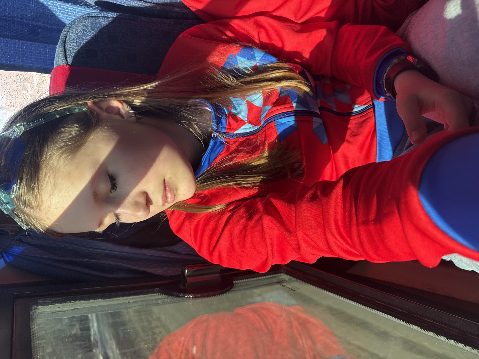 A young girl is seen resting, possibly sleeping, in what appears to be a vehicle. She is wearing a red and blue jacket and a blue headband. Her hand rests on a gray fabric. In the foreground, there is a glass surface, possibly a window or a screen, with a reflection of a red object. The location, Челва, Russia, is not evident in the visual details provided.