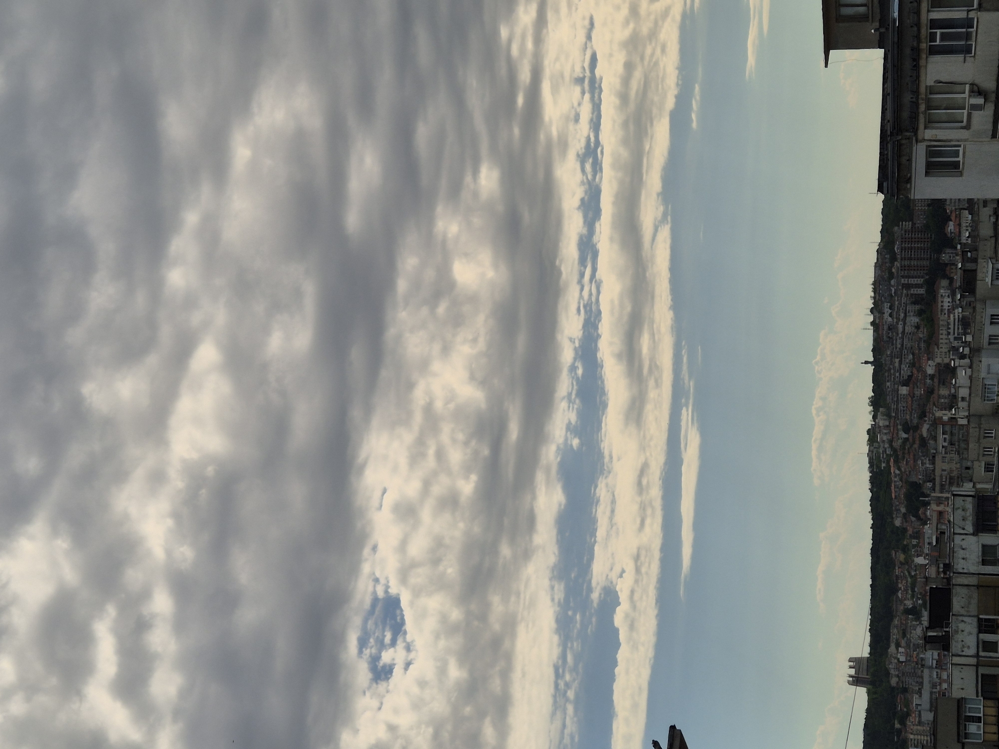The image is a vertical, eye-level shot of a city skyline under a cloudy sky. The foreground features the tops of several buildings, characterized by their muted colors and architectural features like windows and balconies. A portion of a dark, angled structure, possibly a roof or awning, is visible on the left side. The midground displays a dense urban landscape with buildings of varying heights and colors, nestled among trees on a hill. A distinctive, tall, light-colored structure with a stepped roofline stands out on the left side of the hill. In the distance, the sky is filled with layers of clouds, ranging from light, wispy formations to heavier, darker gray masses. A small patch of clear blue sky is visible between the cloud layers. The overall impression is of a city on an overcast day, with a slightly muted color palette.