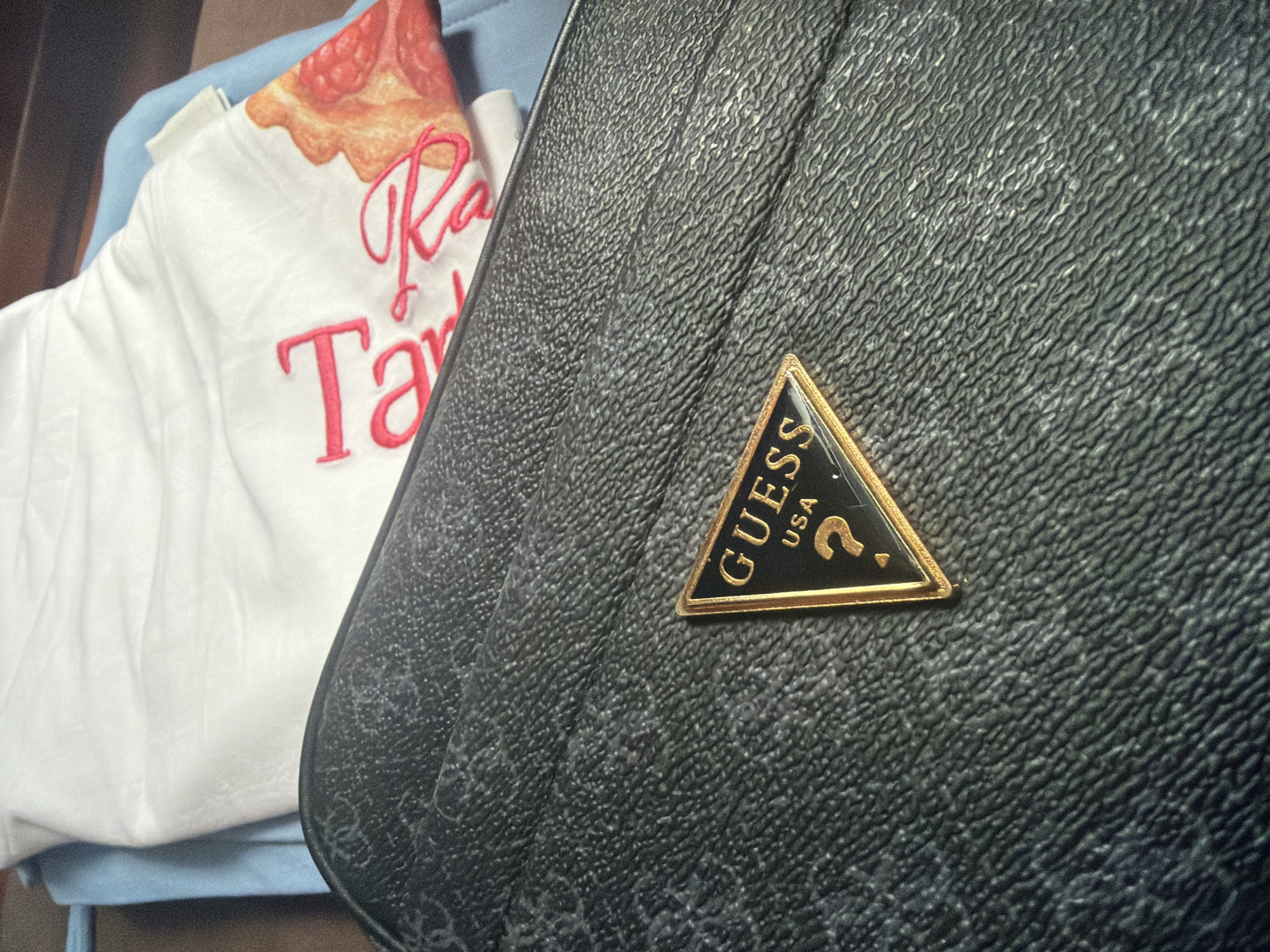 The image shows a close-up of a black textured surface, likely a bag, with a gold and black triangular Guess USA logo on it. A white fabric with red text and an image of pizza is visible in the upper portion of the frame, partially overlapping the black surface. The scene appears to be indoors, with no specific identifiable landmarks to suggest a location in Vratsa, Bulgaria.