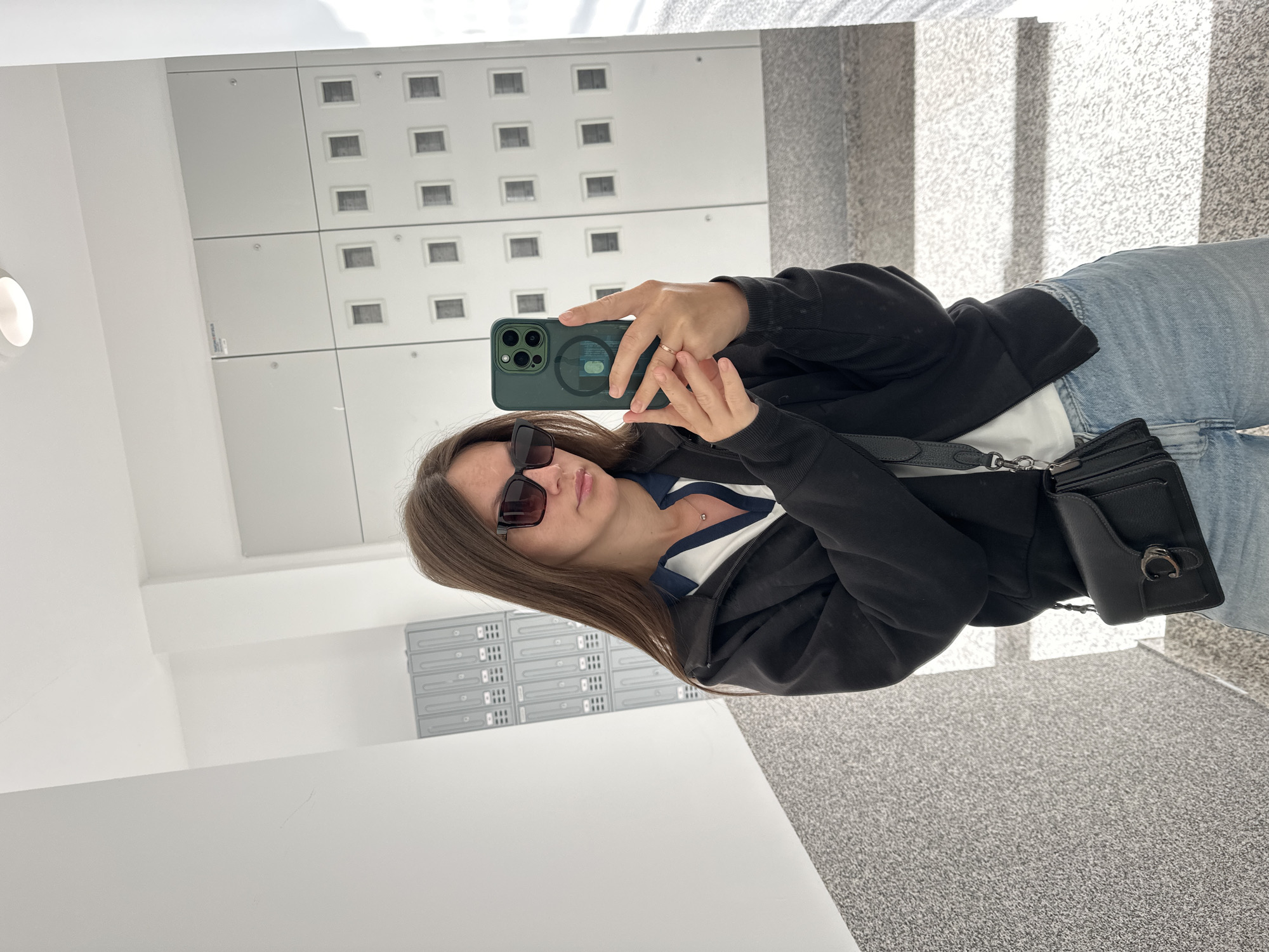 A person stands in a mirrored space, possibly an indoor area, and takes a selfie with a green iPhone. The individual wears sunglasses, a black zip-up jacket over a white shirt with a dark blue collar, and blue jeans. A black purse hangs off to the side of their body. The background includes a white wall with a grid pattern of small squares and a floor that appears to be carpeted. A separate wall with a locker or cabinet system is partially visible in the reflection. The location is assumed to be in Donji Pobori, Montenegro.