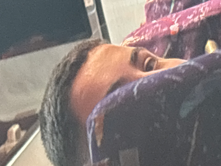 The image depicts a partial view of an individual within an interior setting, likely a vehicle, in Aksakovo, Bulgaria. The central subject is an adult with dark, short-cropped hair, whose forehead and right eye are visible. The skin tone is warm, appearing tan or olive. The person's gaze is directed towards the lower right. The left side of the individual's face and body is obscured by a dark blue or purple patterned fabric, which extends into the foreground. To the right of the individual, a pink or magenta patterned fabric is present, partially obscuring the right side of the body. In the upper background, a horizontal light-colored line is visible, consistent with a window frame or structural division. Above and to the right of this line, a dark, reflective surface, possibly a window, shows a faint reflection. The overall scene suggests the individual is seated or resting within an enclosed space.